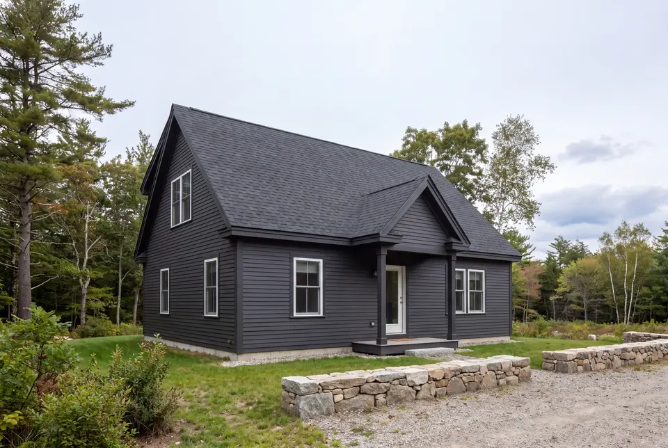 custom home builder in South Portland, ME: illustrative scene, A dark charcoal cottage with a gabled roof and white-trimmed windows, set in a natural landscape with trees and a gravel driveway