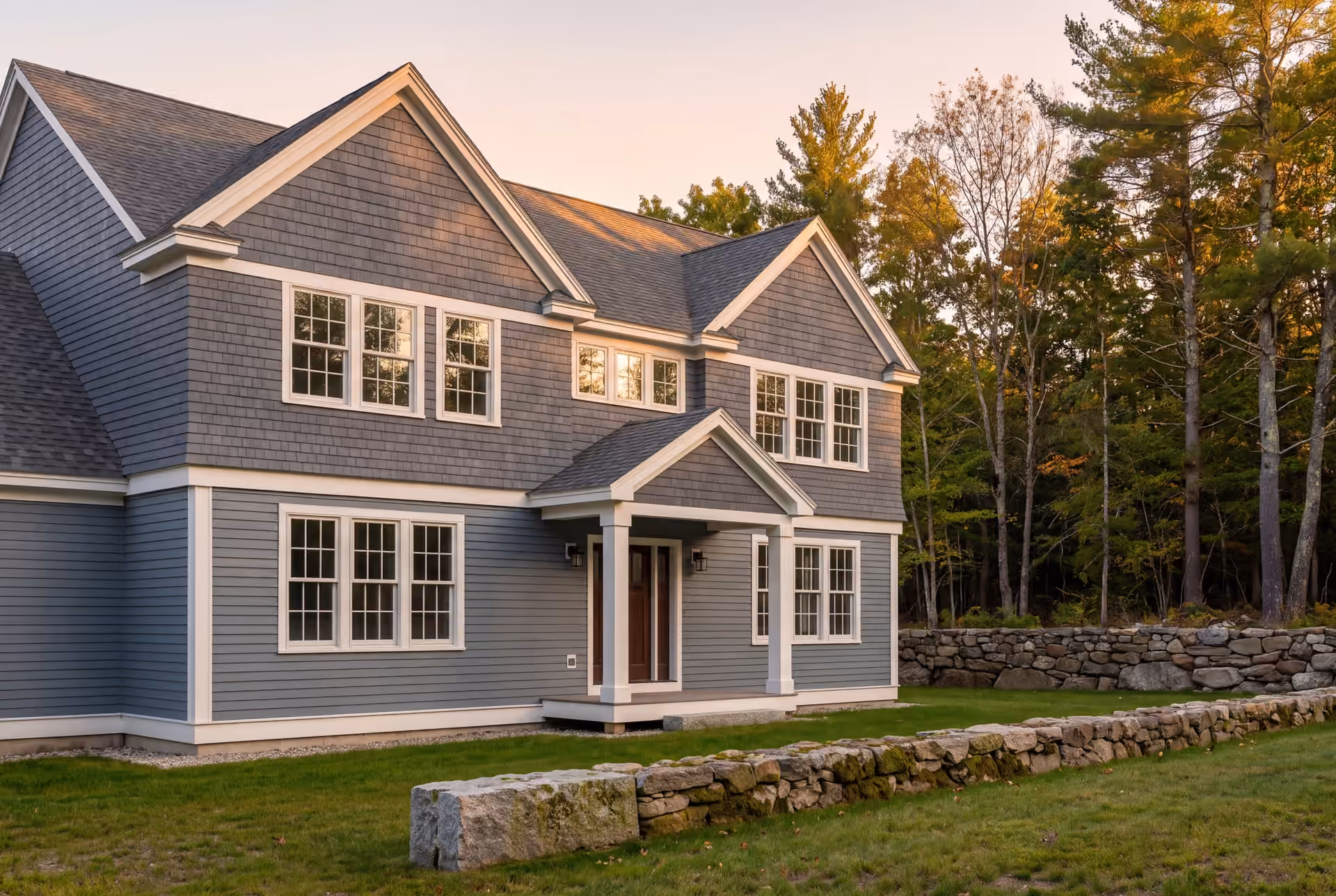 custom home builder in Standish, ME: illustrative scene, A newly built two-story home with blue-gray siding and a dark roof, seen from a three-quarter angle with a stone wall and trees in the background