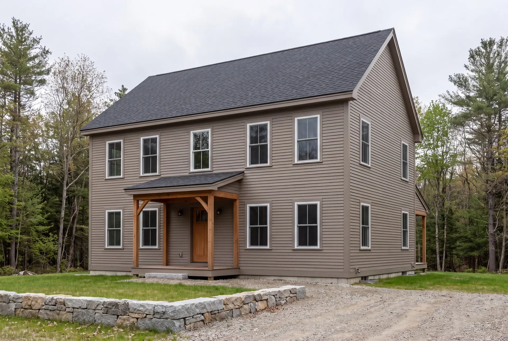 Illustration: A newly built two-story farmhouse with taupe siding, a dark gray roof, and white-trimmed windows, viewed from a three-quarter angle with a gravel driveway and stone wall in the foreground