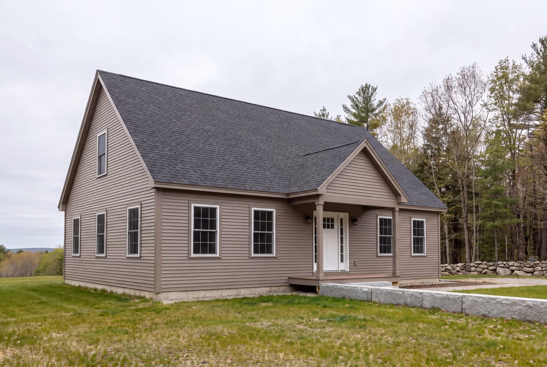 custom home builder in Westbrook, ME: illustrative scene, A newly built single-story house with taupe siding, a dark gray roof, and a covered front porch, surrounded by a green lawn and trees