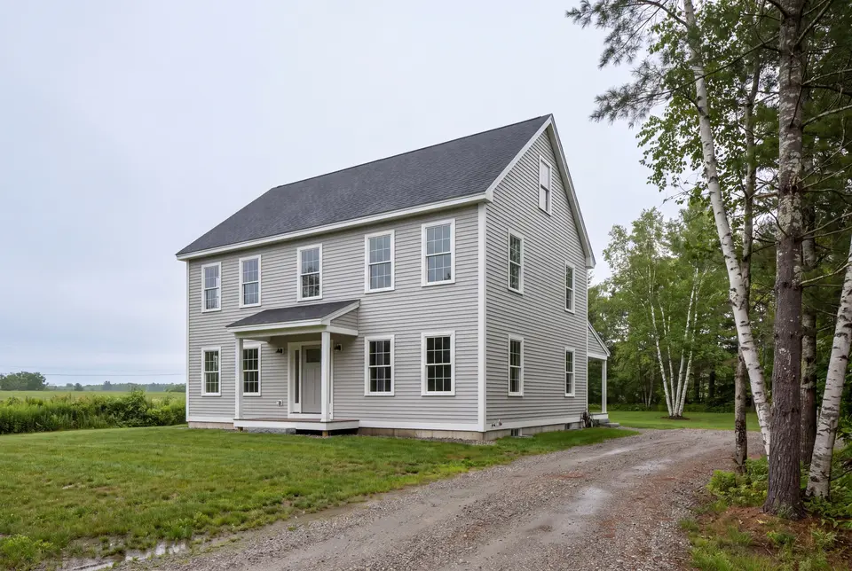 Illustration: A newly built two-story Colonial house with light gray siding and a dark gray roof, viewed from a three-quarter angle with a gravel driveway and trees in the foreground