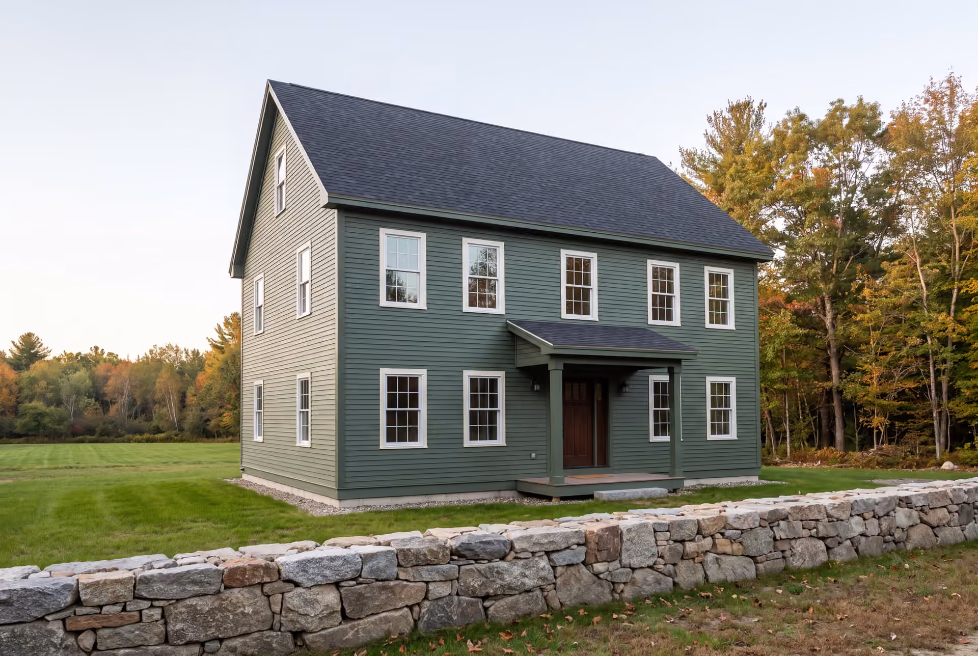 custom home builder in Windham, ME: illustrative scene, A green farmhouse with a dark roof and white windows, seen from a three-quarter angle, with a stone wall in the foreground and autumn trees in the background