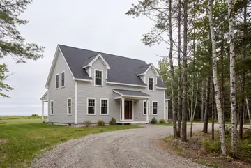 custom home builder: illustrative scene, A newly built Cape house with light gray siding and a dark gray roof, featuring two dormers and a covered front entry, set in a grassy area with a gravel driveway and trees