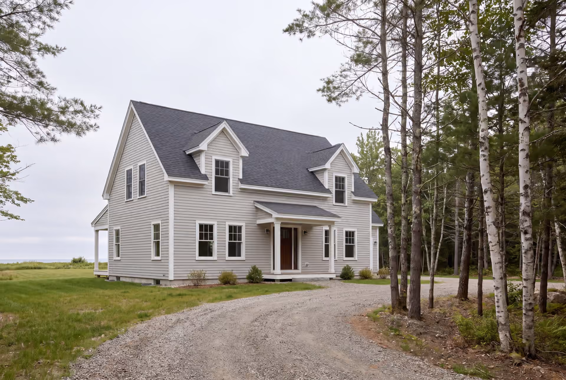 custom home builder: illustrative scene, A newly built Cape house with light gray siding and a dark gray roof, featuring two dormers and a covered front entry, set in a grassy area with a gravel driveway and trees