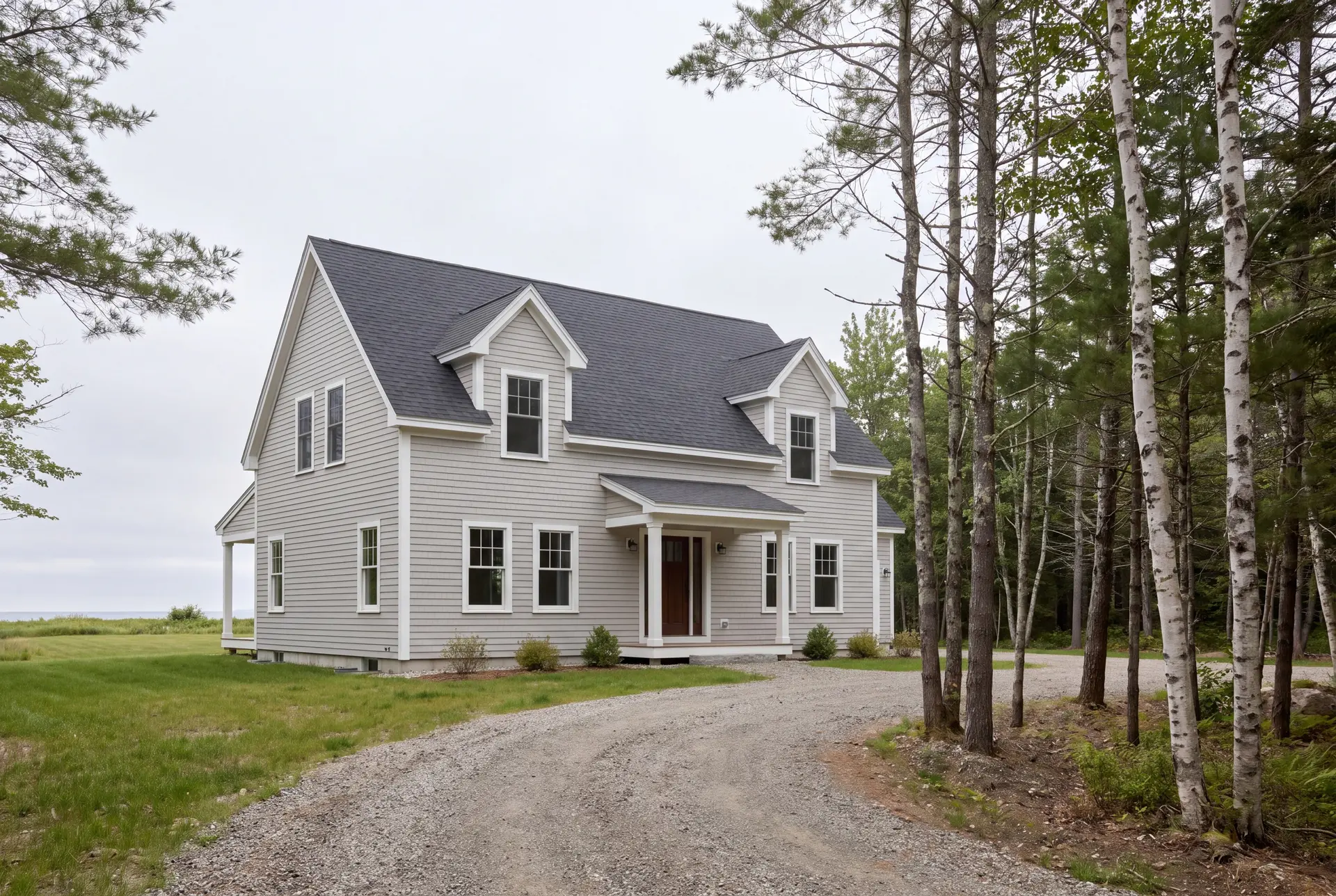custom home builder: illustrative scene, A newly built Cape house with light gray siding and a dark gray roof, featuring two dormers and a covered front entry, set in a grassy area with a gravel driveway and trees
