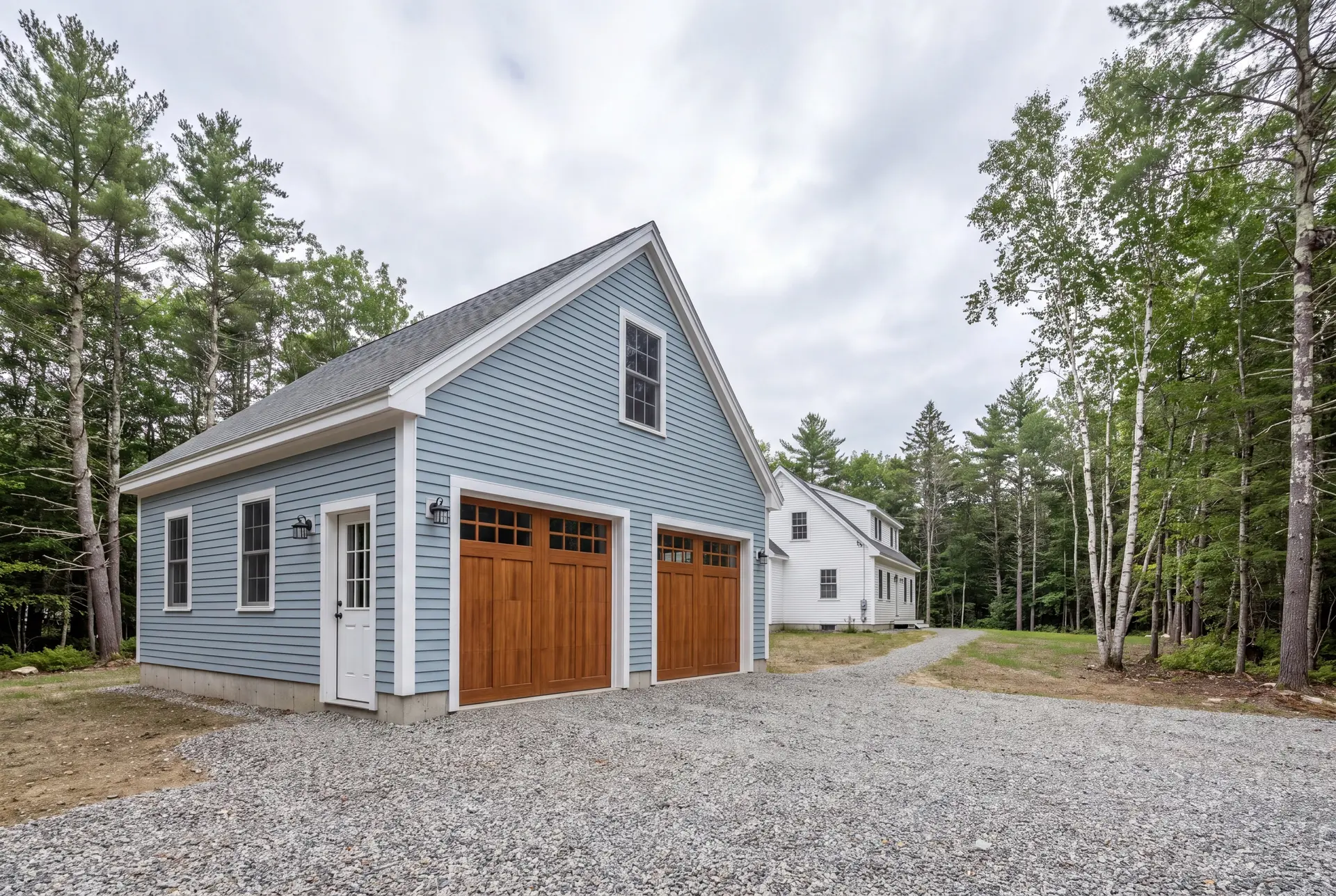 Illustration: A newly built light blue garage with two wooden garage doors and a gravel driveway, next to a white house and surrounded by trees