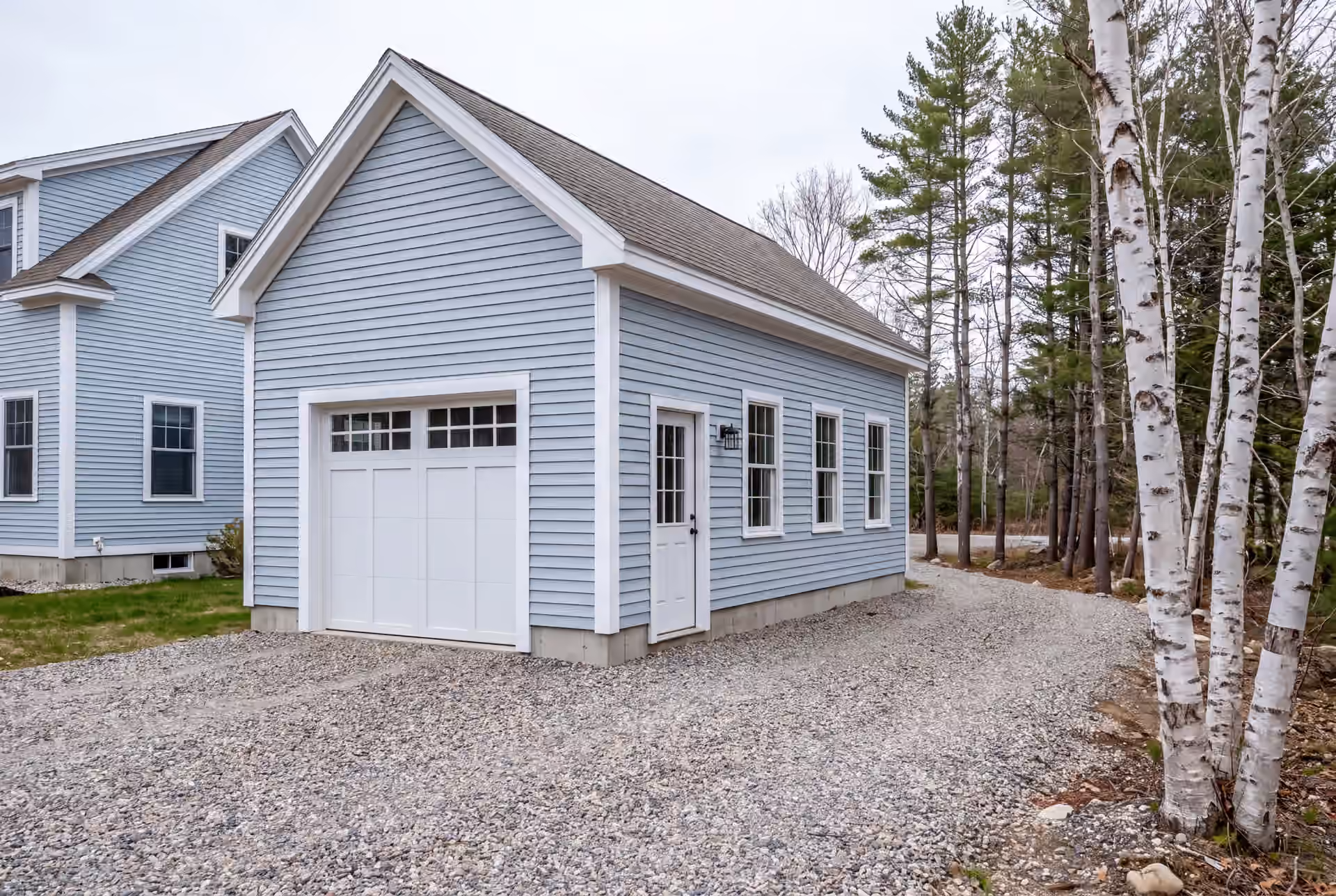 garage builder in Biddeford, ME: illustrative scene, A newly built light blue garage with a white garage door and service door, next to a matching house, with a gravel driveway and trees