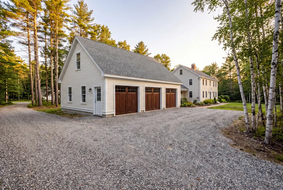 Illustration: A newly built three-bay garage with white siding and dark wood doors, next to a house, with a gravel driveway and trees