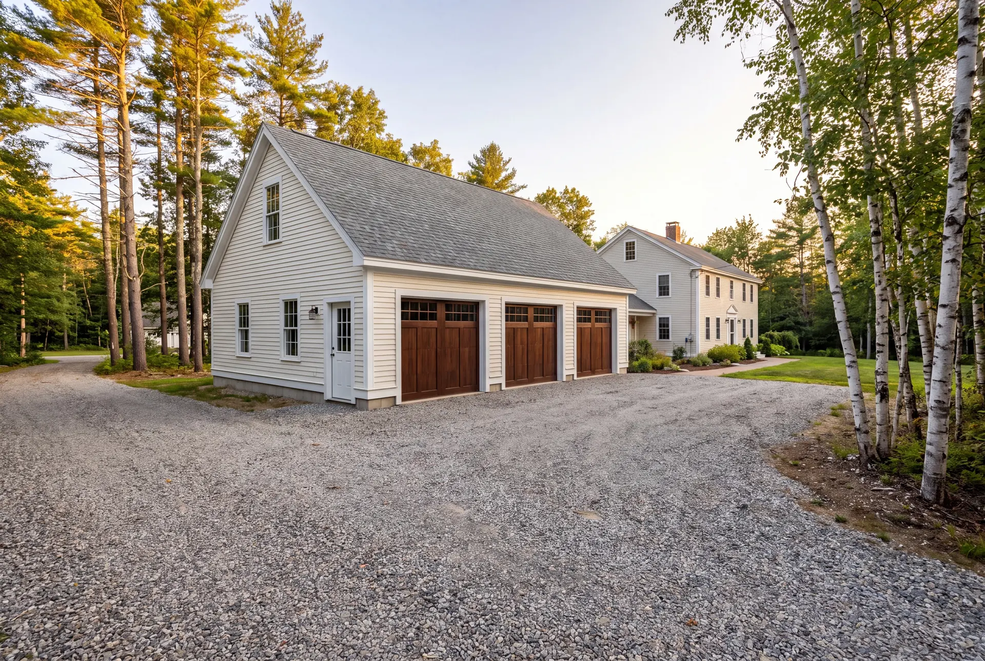 Illustration: A newly built three-bay garage with white siding and dark wood doors, next to a house, with a gravel driveway and trees