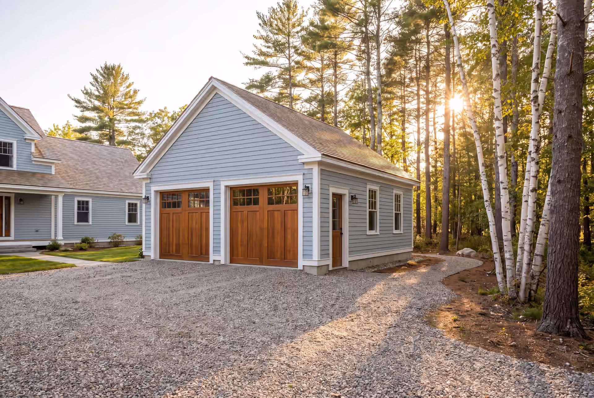 garage builder in Buxton, ME: illustrative scene, A light blue detached garage with two wooden garage doors and a gravel driveway, set against a backdrop of tall trees and a partially visible house
