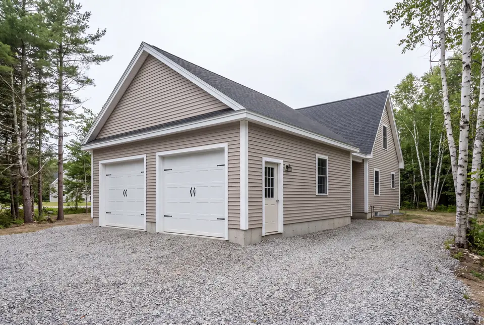 Illustration: A newly built two-bay garage with mushroom-colored siding and a dark roof, attached to a house, with a gravel driveway and surrounding trees