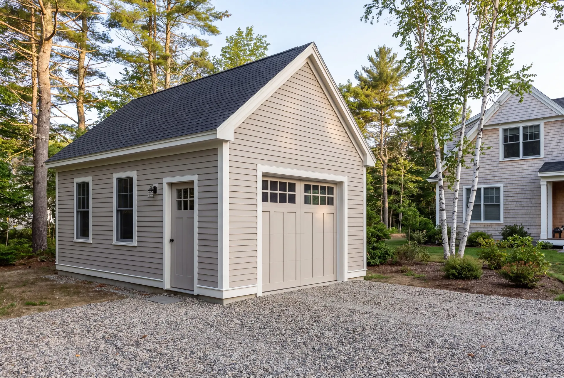 Illustration: A newly built, light-colored residential garage with a dark roof and gravel driveway, situated next to a house with trees in the background