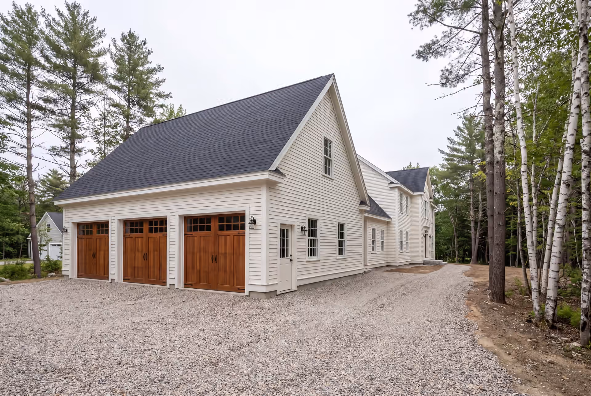 garage builder in Cape Elizabeth, ME: illustrative scene, A newly built white residential garage with three wooden doors and a gravel driveway, attached to a matching house, surrounded by trees under a cloudy sky