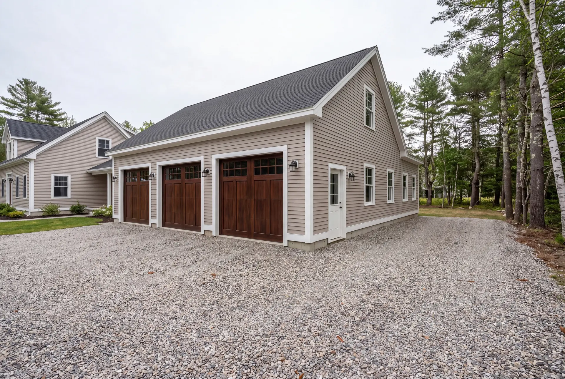 Illustration: A newly built three-bay garage with mushroom-colored siding and dark brown doors, next to a matching house, with a gravel driveway and surrounding trees under an overcast sky