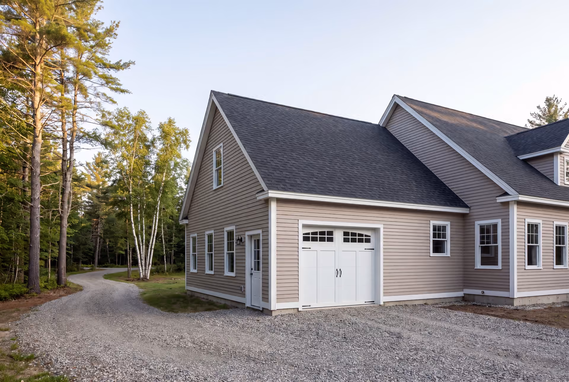 garage builder in Cumberland, ME: illustrative scene, A newly built, single-bay garage with mushroom-colored siding and a dark roof, attached to a house, with a gravel driveway and trees