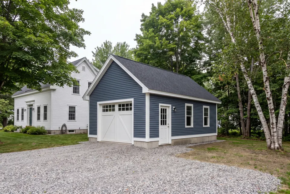 Illustration: A newly built blue garage with a white garage door and service door, next to a white house, with a gravel driveway and trees