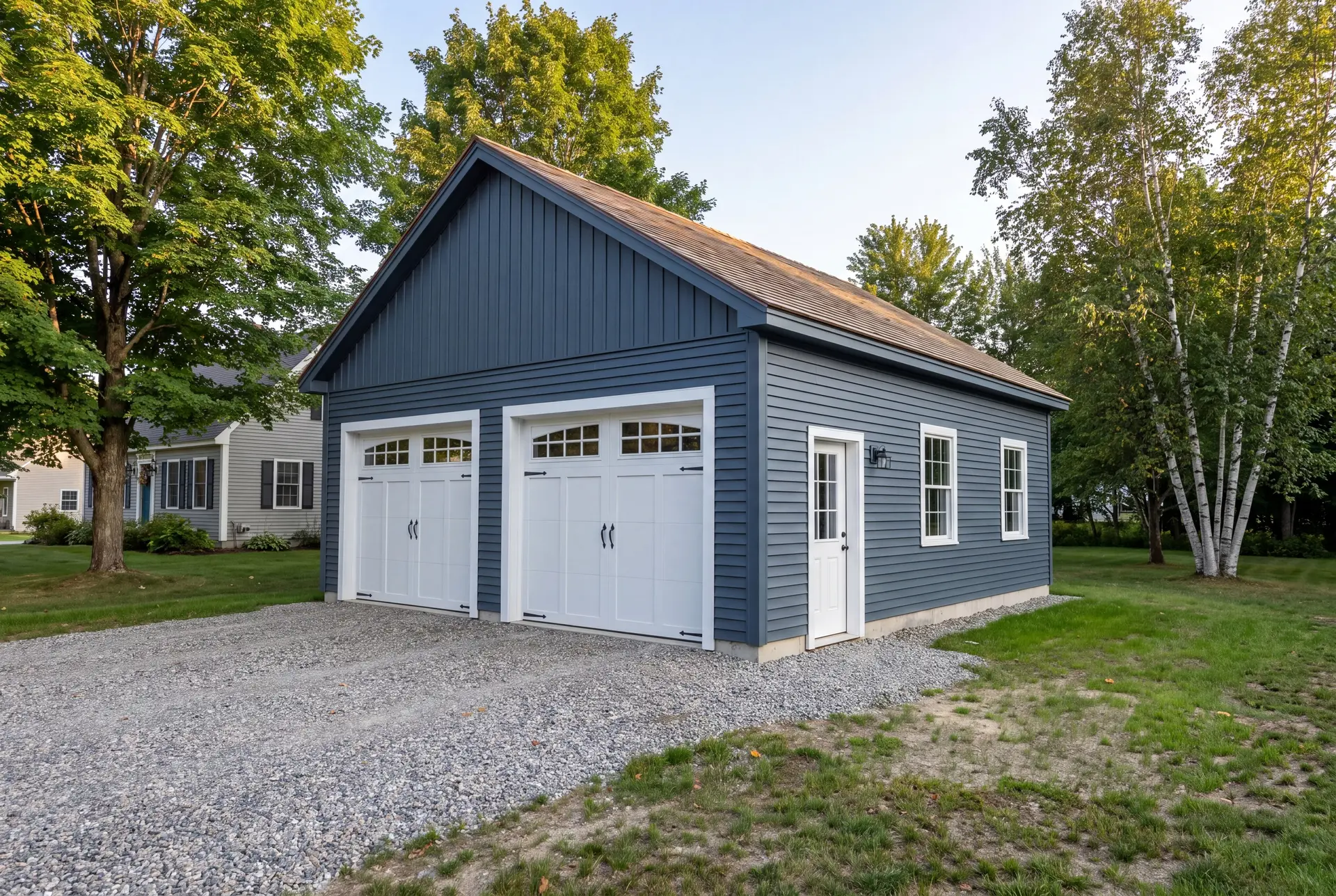 Illustration: A newly built, detached two-bay garage with deep slate blue siding, two white carriage-style doors, and a gravel driveway