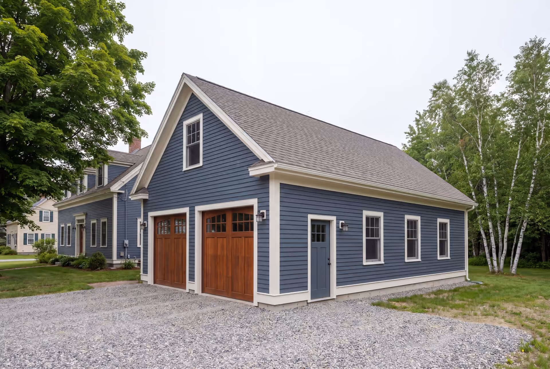 garage builder in Dayton, ME: illustrative scene, A newly built, detached two-bay garage with deep slate blue siding and wooden carriage-style doors, next to a house