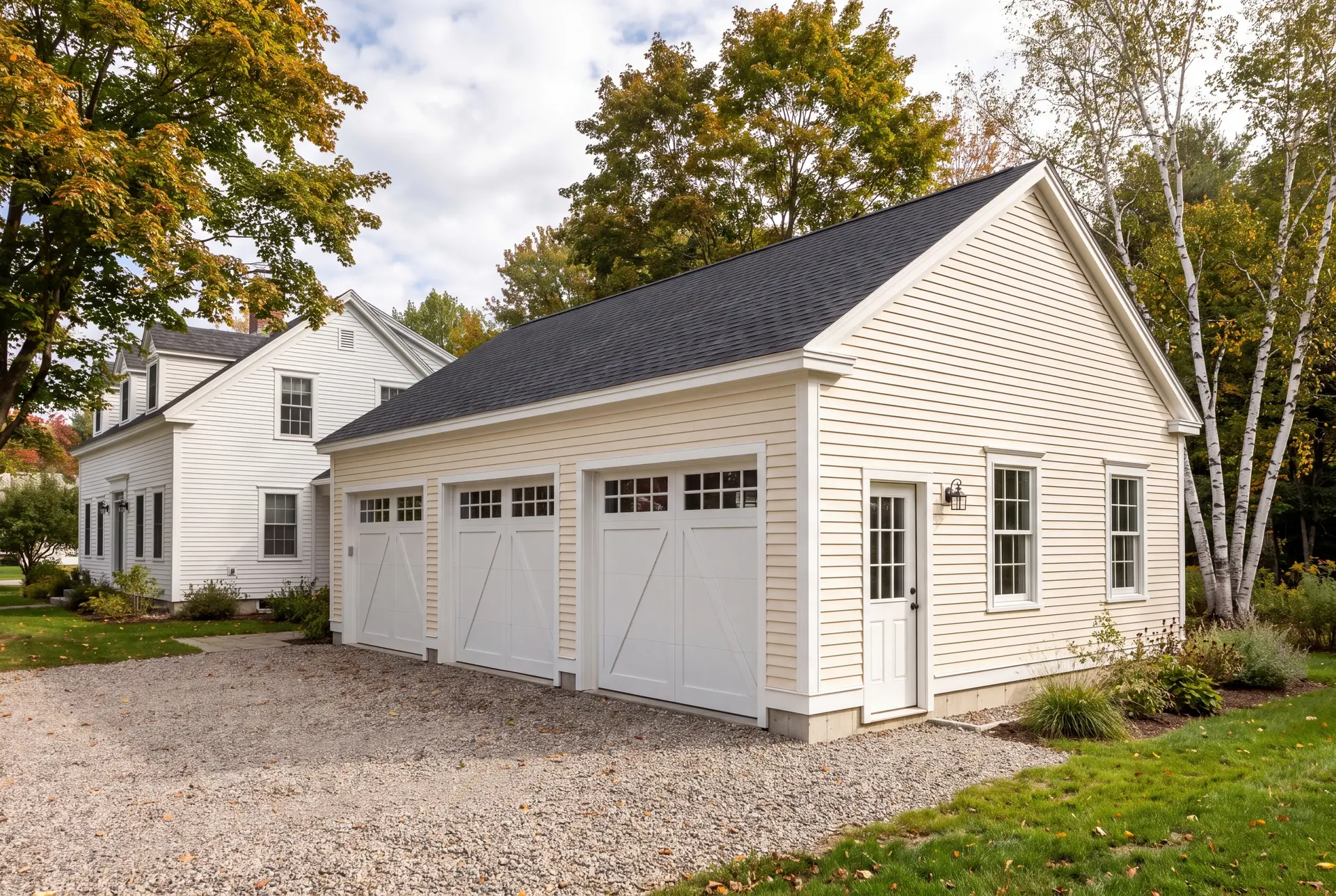 Illustration: A newly built, detached three-car garage with ivory siding and a dark roof, situated next to a white house, with a gravel driveway and autumn trees