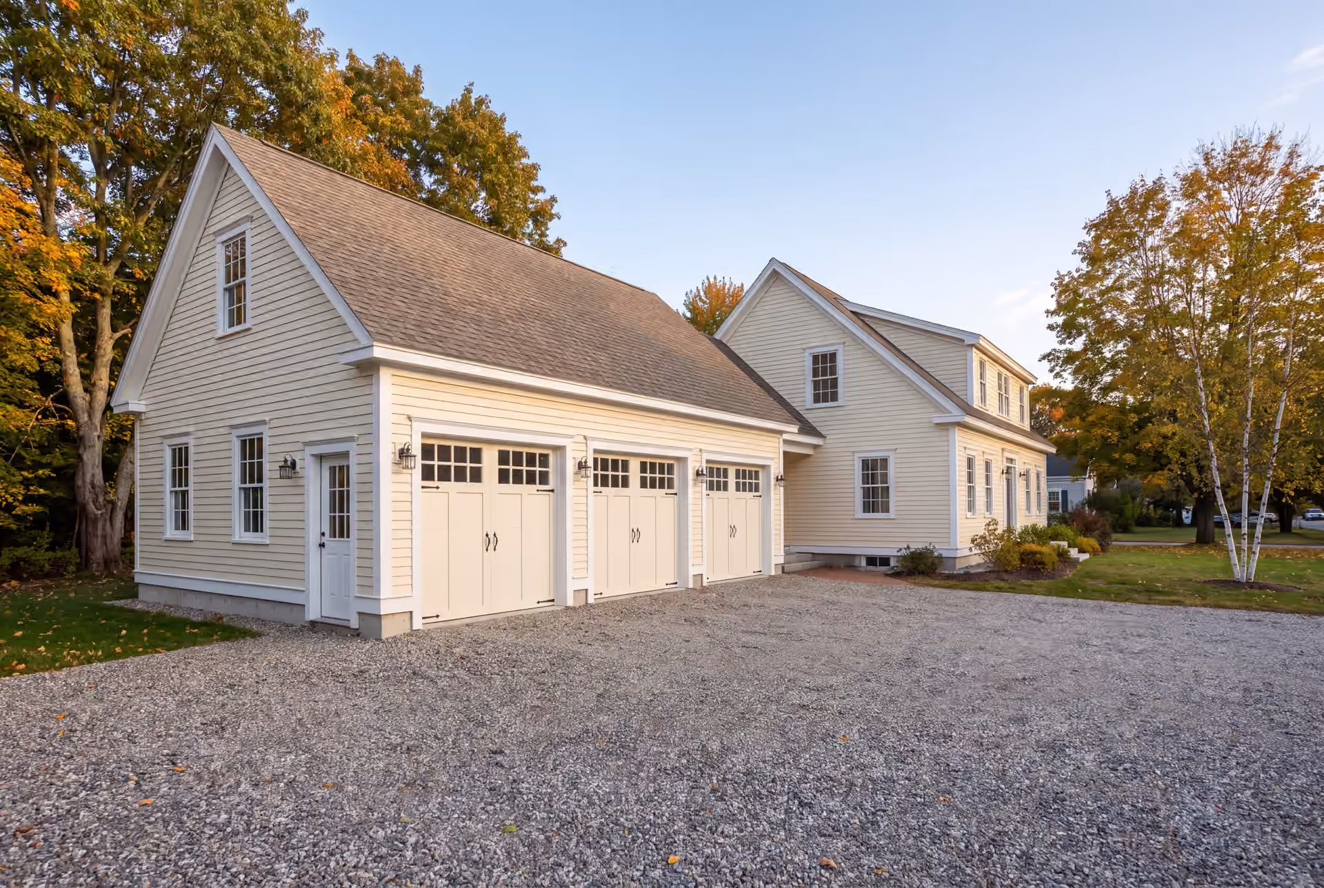 garage builder in Falmouth, ME: illustrative scene, A newly built, light-colored three-bay garage with carriage doors and a matching house, set against a backdrop of autumn trees and a gravel driveway