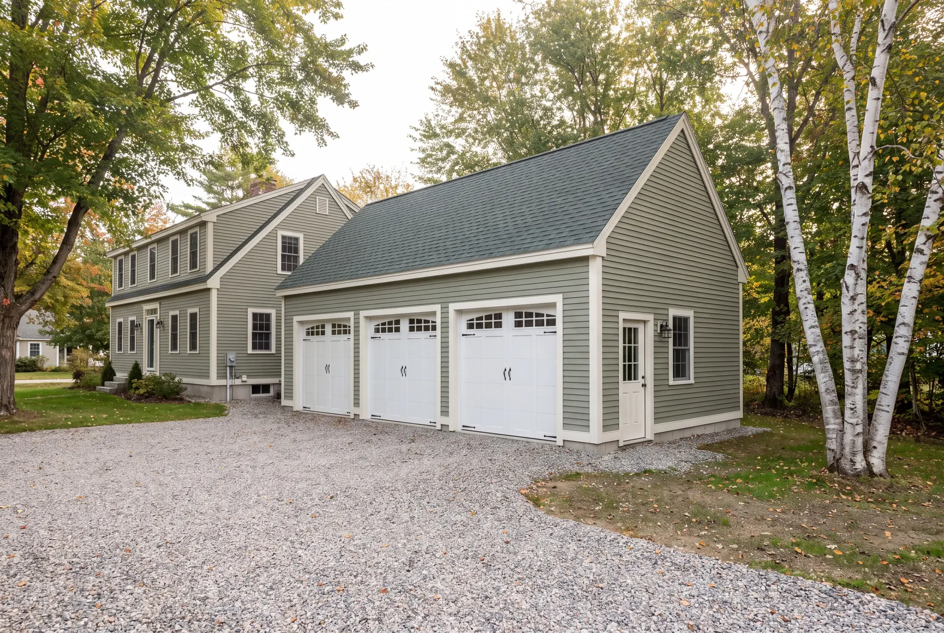 Illustration: A newly built, detached three-bay garage with sage green siding and a dark green roof, next to a matching house, with a gravel driveway and trees