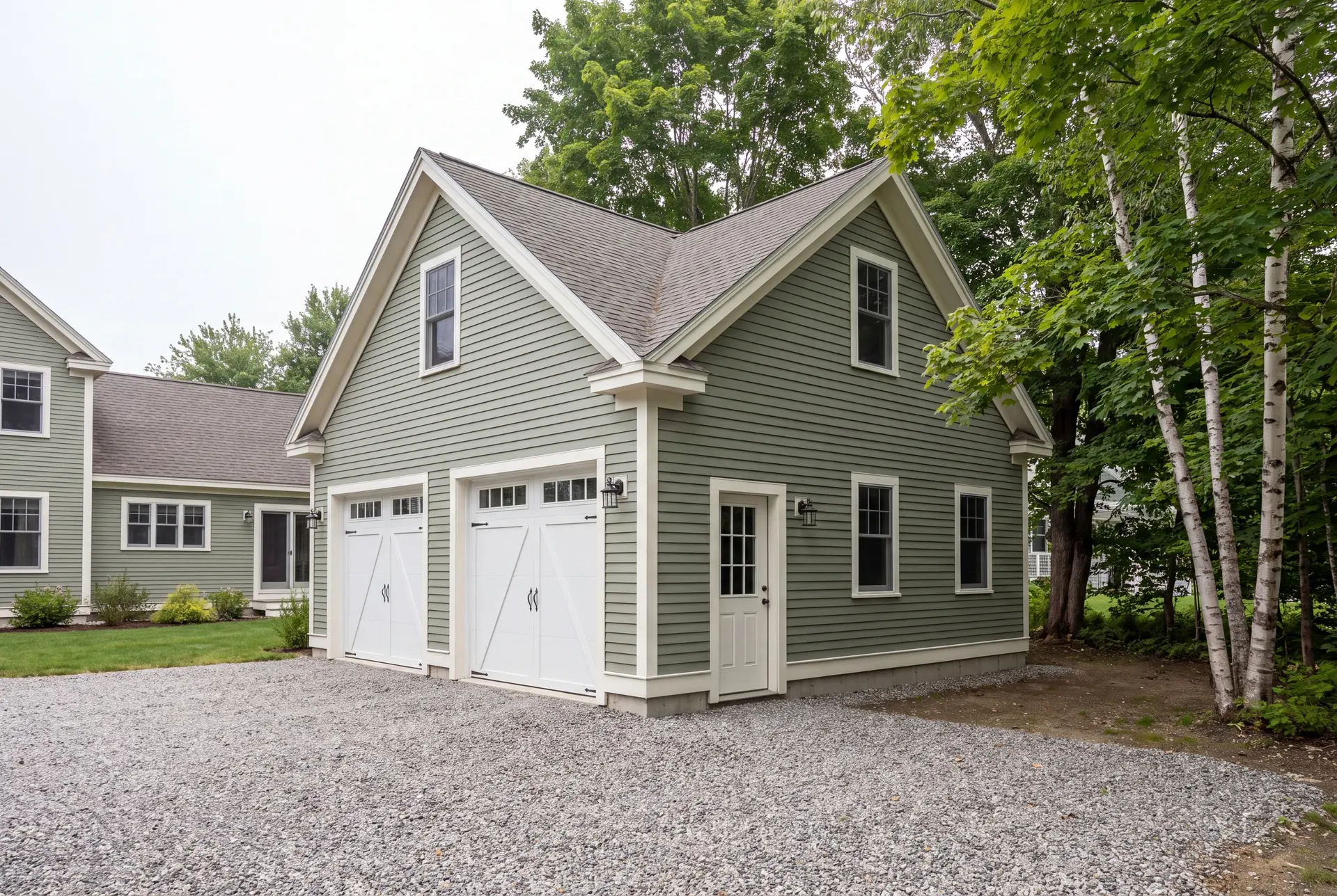 Illustration: A newly built sage green two-car garage with white trim and carriage doors, next to a matching house, with a gravel driveway and surrounding trees