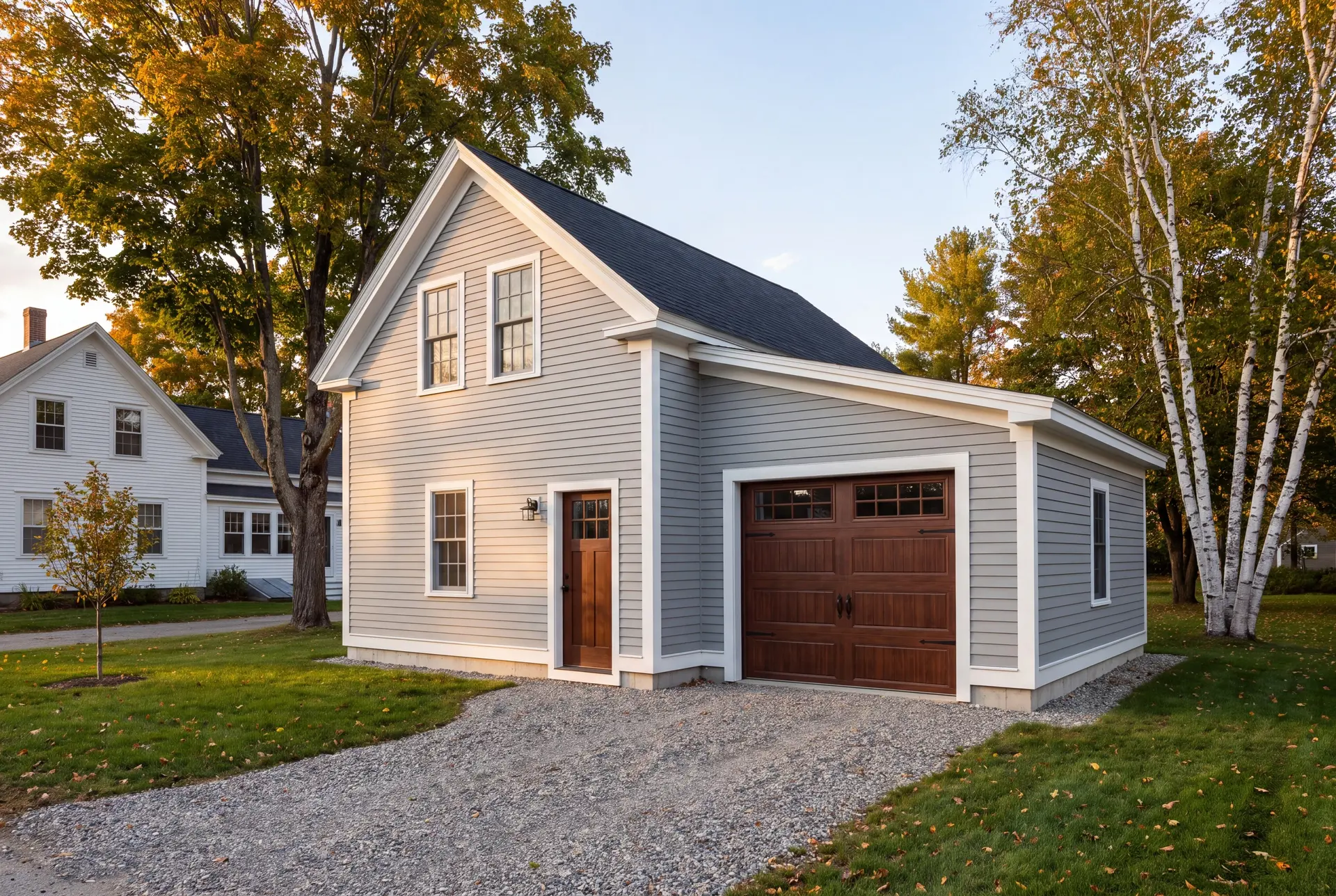 Illustration: A newly built gray garage with a brown carriage door and service door, next to a white house, surrounded by trees and a gravel driveway