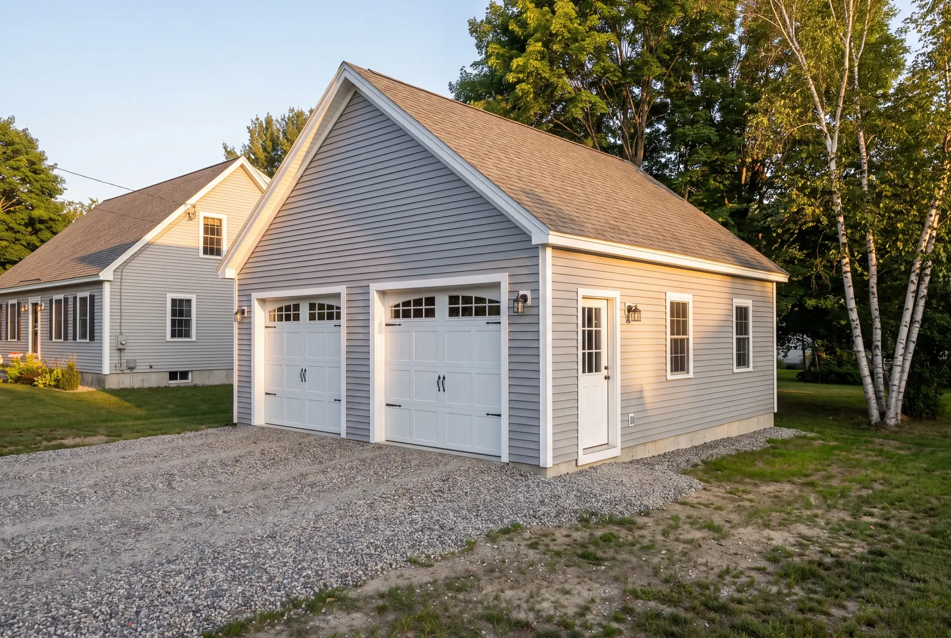 Illustration: A newly constructed light gray garage with two white carriage doors and a service door, next to a house, with a gravel driveway and green landscaping