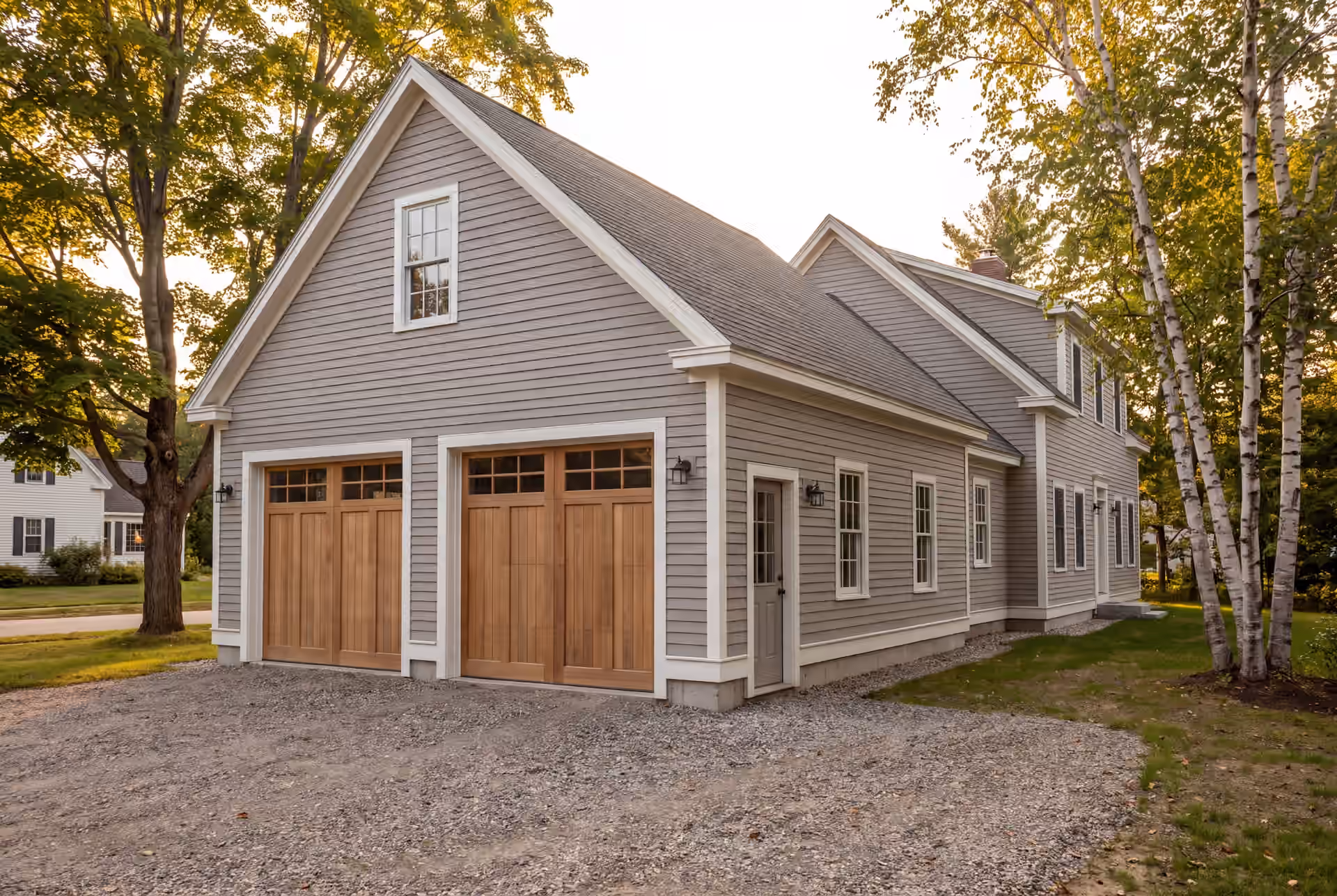 garage builder in Gorham, ME: illustrative scene, A newly built two-bay garage with light gray siding and wooden carriage doors, attached to a matching house, with a gravel driveway and surrounding trees