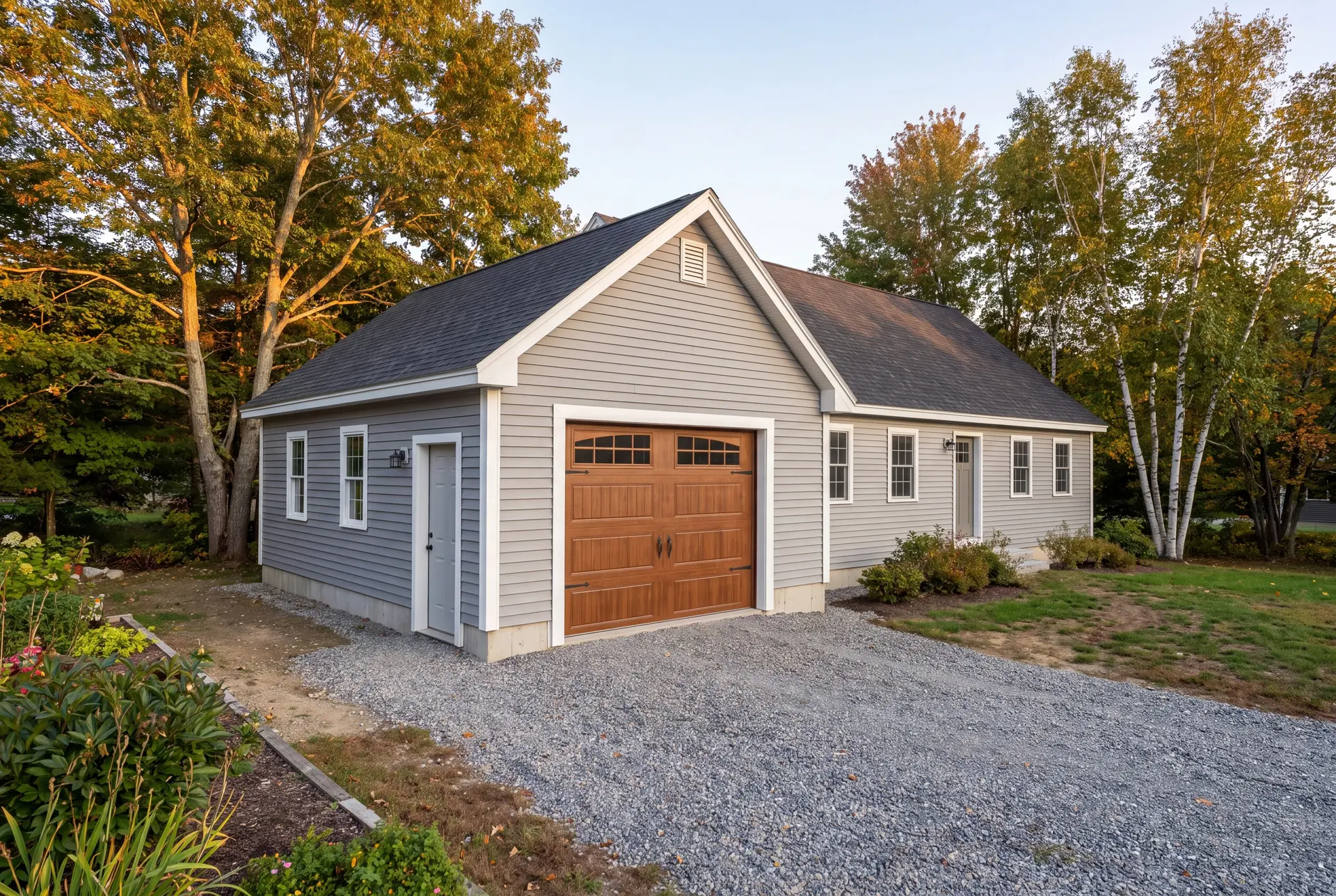 Illustration: A newly built gray garage with a brown garage door and gravel driveway, surrounded by trees and landscaping