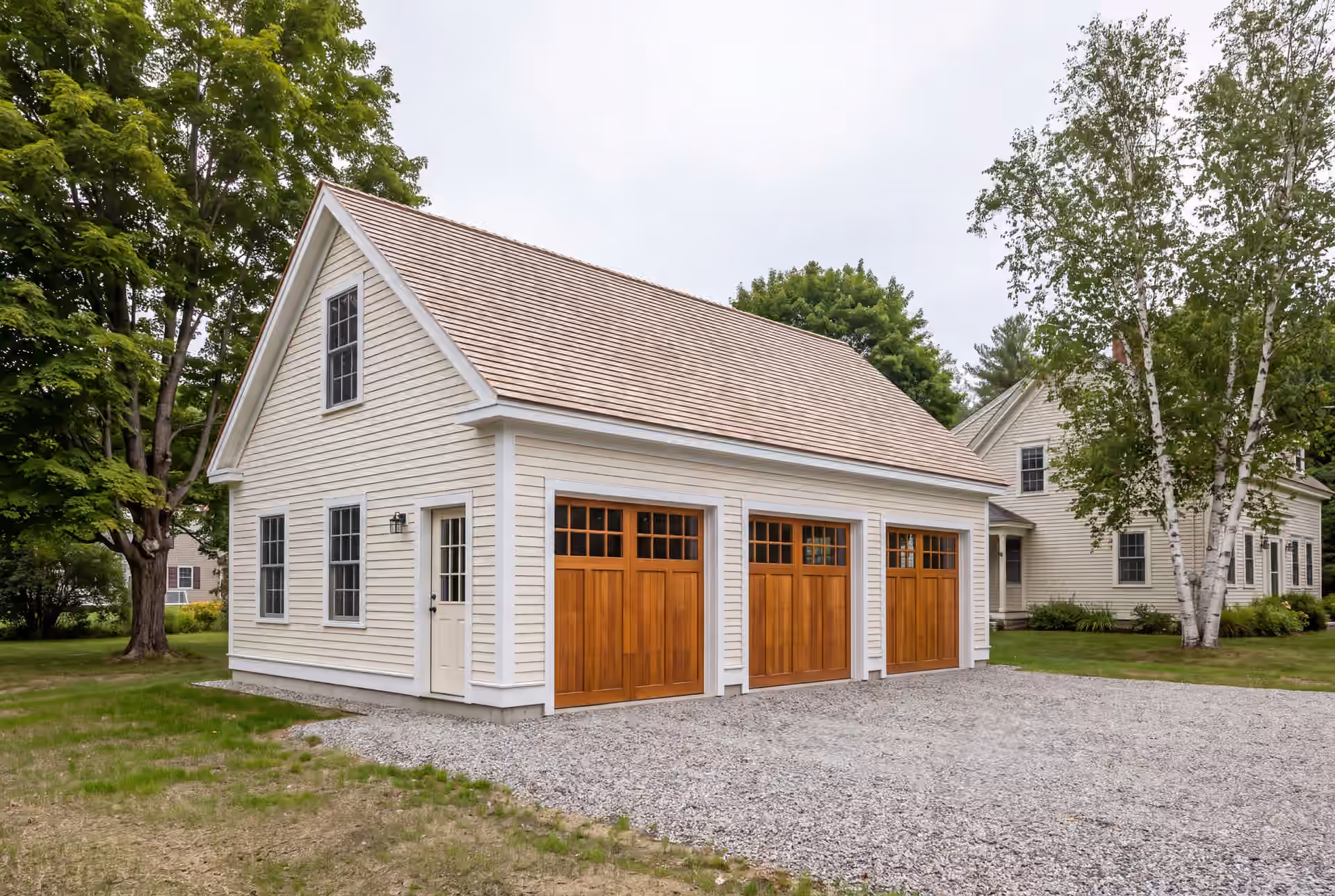 garage builder in Gray, ME: illustrative scene, A newly built, detached three-bay garage with pale cream siding and a shingle roof, featuring three wooden garage doors and a gravel driveway
