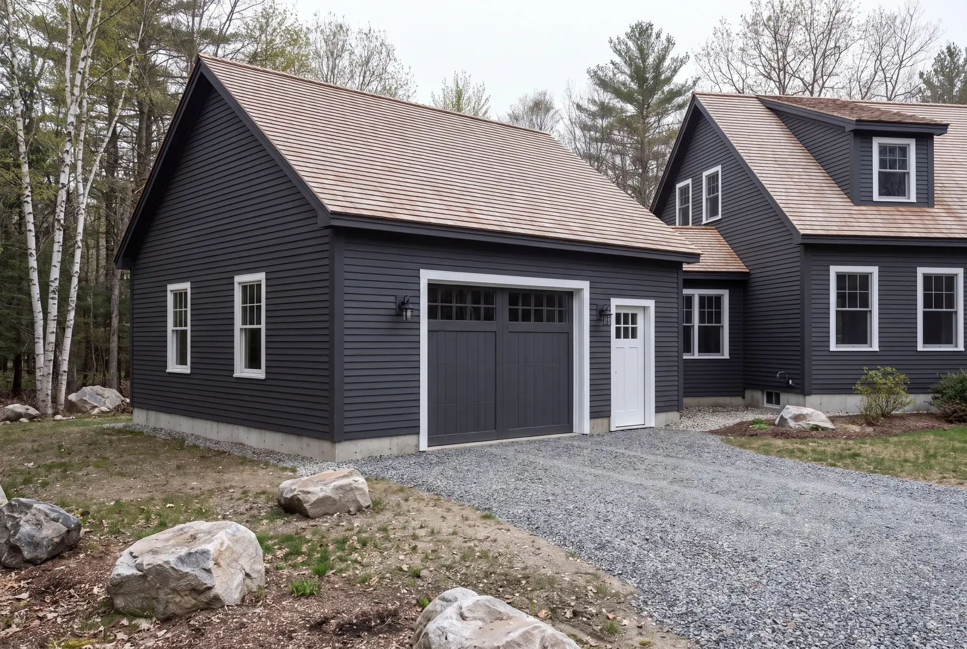 Illustration: A newly built, dark gray clapboard garage with a light brown shingle roof and gravel driveway, next to a matching house