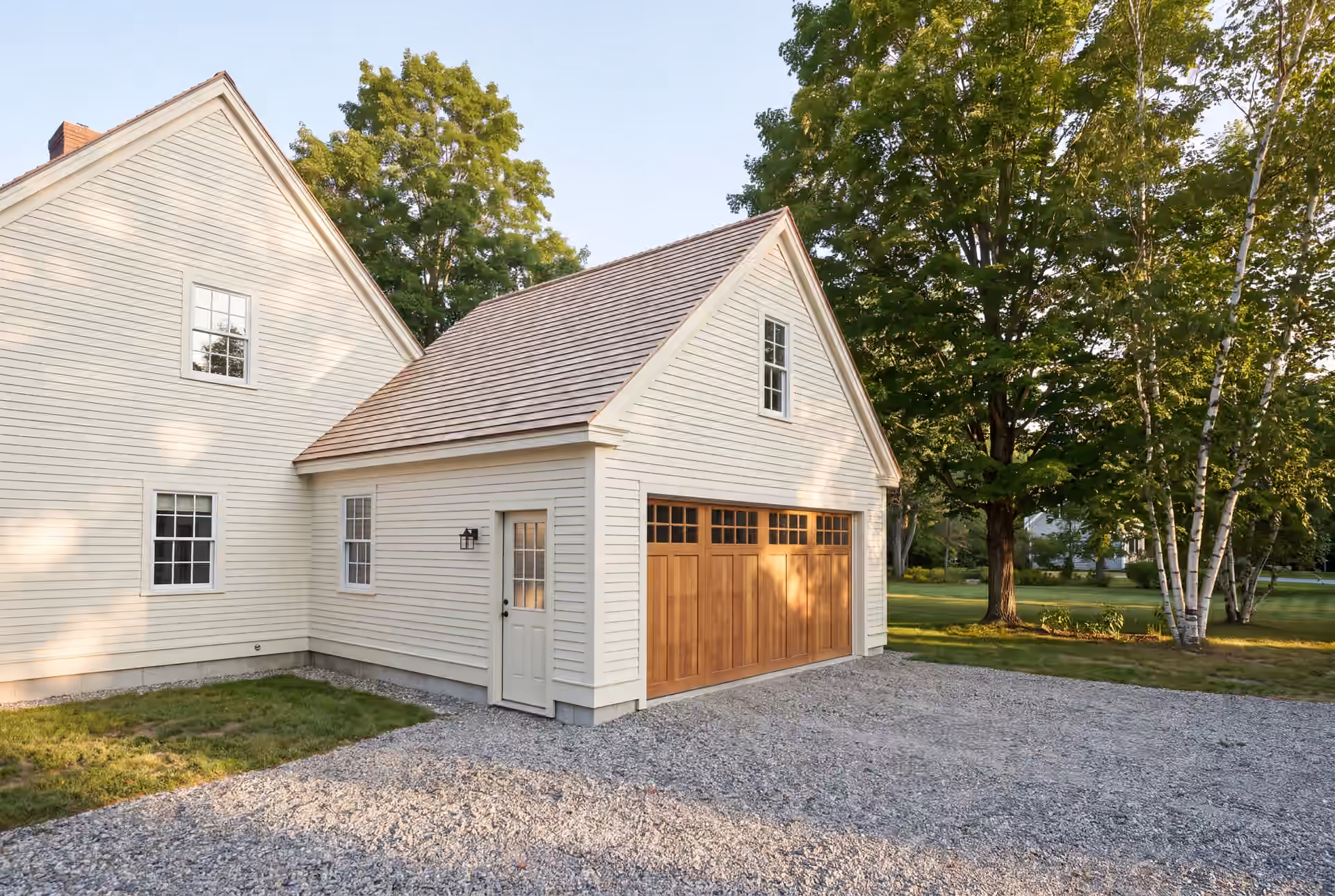 garage builder in Hollis, ME: illustrative scene, A newly built, cream-colored clapboard garage with a wooden carriage door and shingle roof, attached to a matching house, with a gravel driveway and green lawn