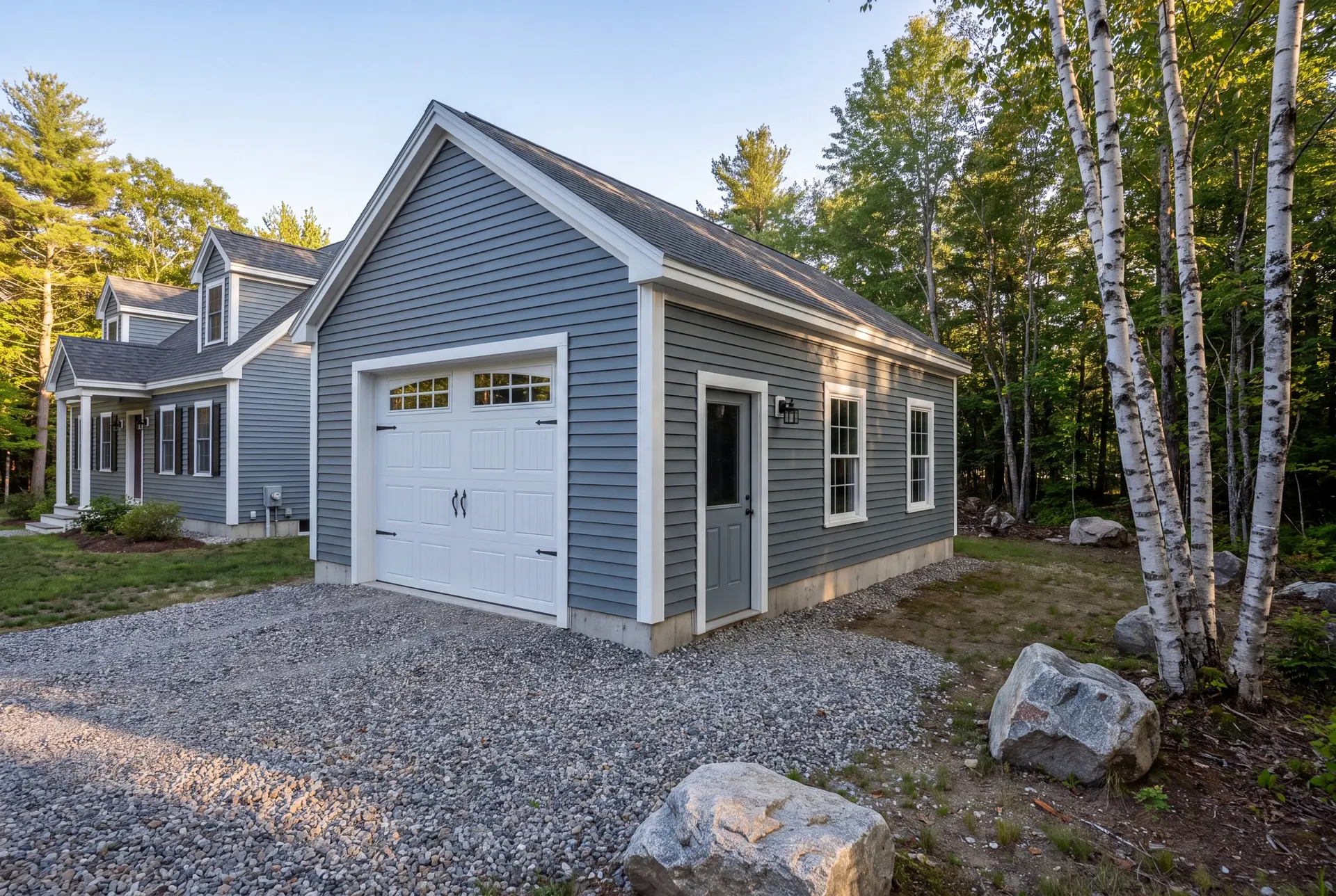 Illustration: A newly built blue-gray garage with a white carriage-style door and a gray service door, next to a house, with a gravel driveway and trees