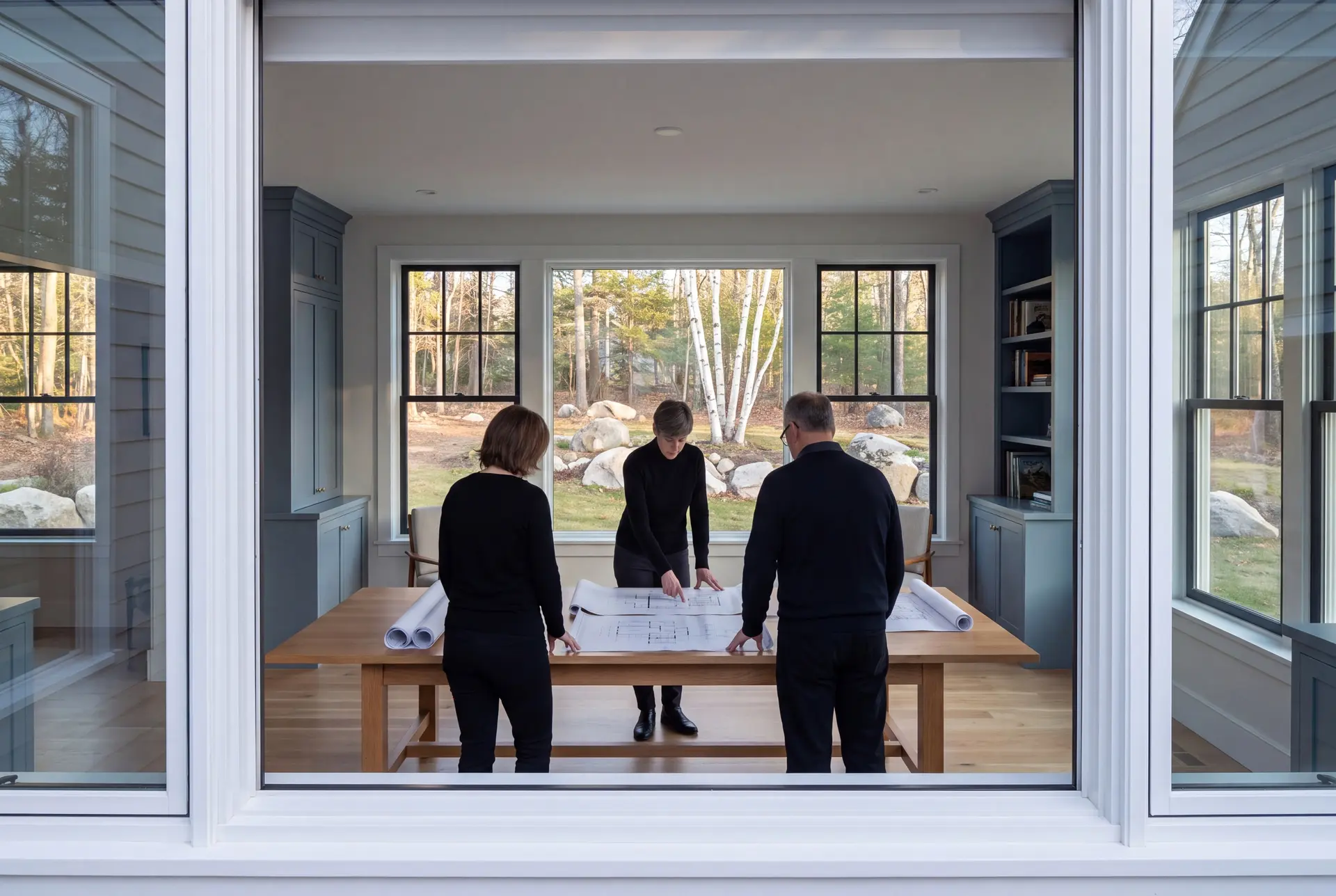 Illustration: Three people, two women and one man, reviewing architectural drawings on an oak table in a modern living room with blue-gray cabinets and large windows