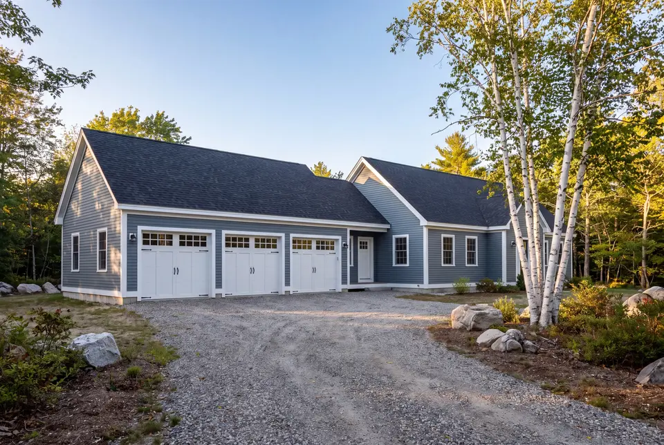 Illustration: A newly built blue-gray house with an attached three-car garage and a gravel driveway, surrounded by trees and rocks under a clear sky