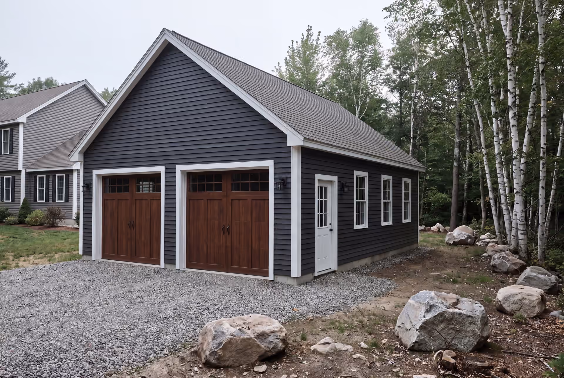 garage builder in Kennebunk, ME: illustrative scene, A newly built dark gray garage with two brown doors and white trim, next to a house and a forest