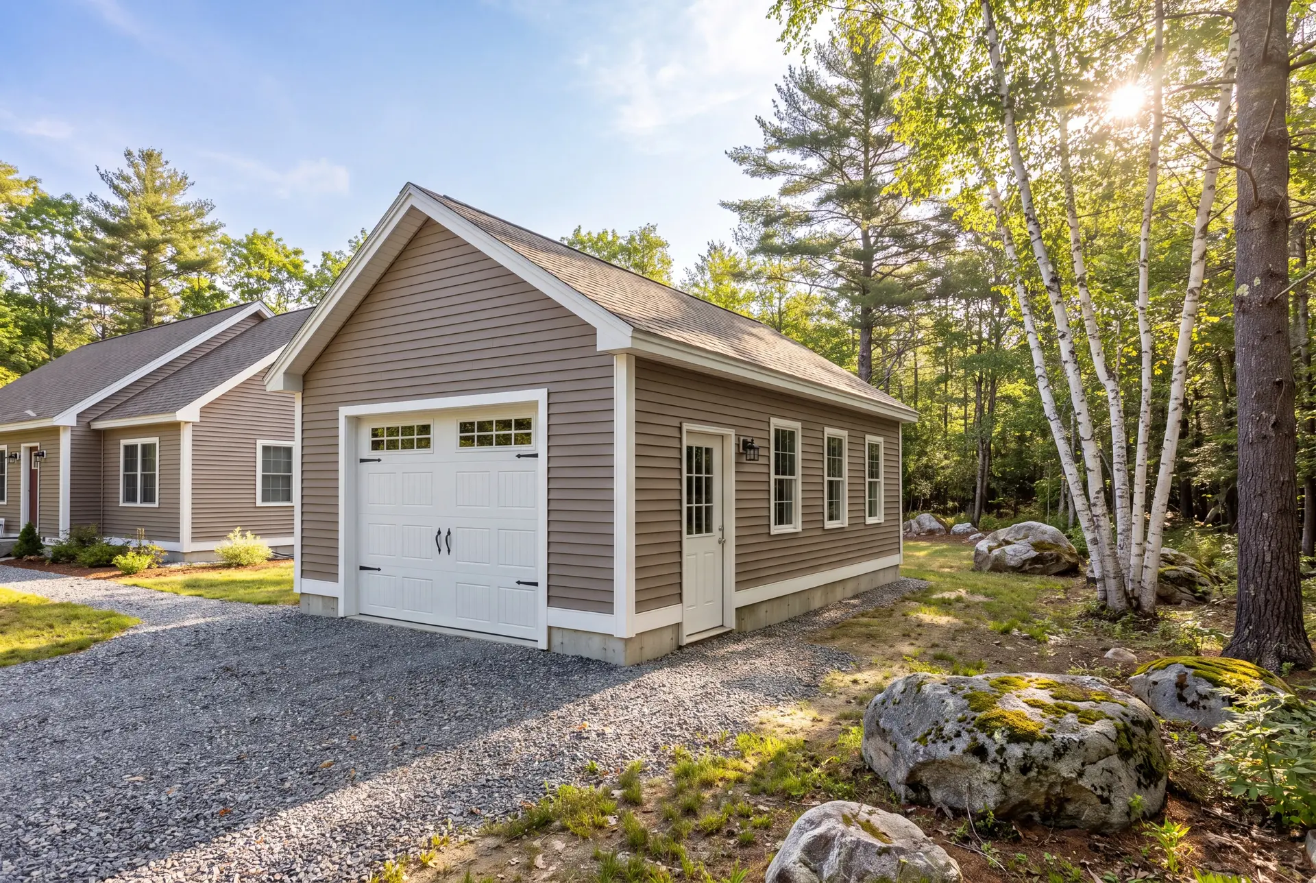 Illustration: A newly built, detached single-bay garage with taupe siding and a white carriage-style door, next to a house, surrounded by trees and rocks