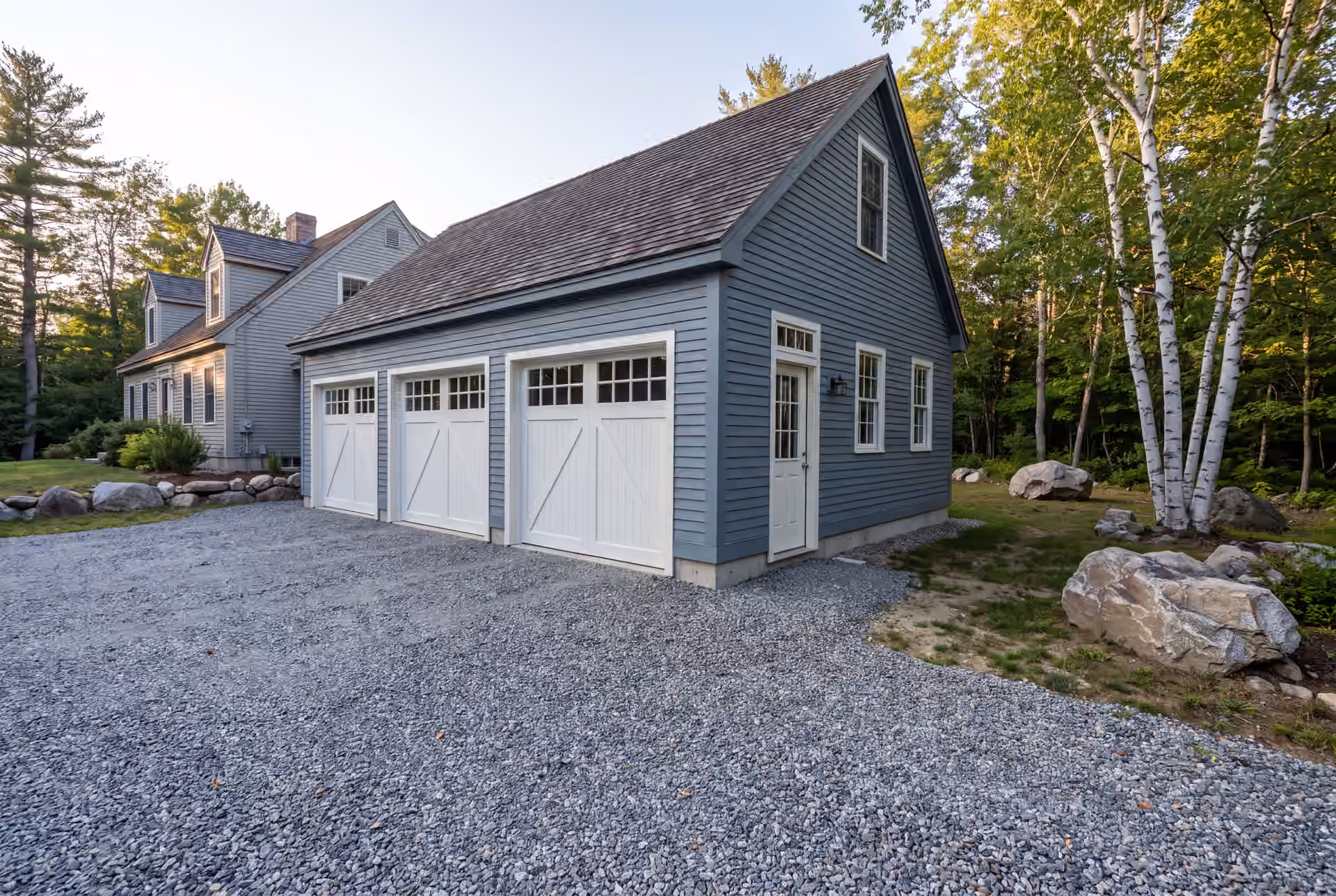 garage builder in Lyman, ME: illustrative scene, A newly built three-bay garage with blue-gray siding and white doors, next to a house, with a gravel driveway and trees