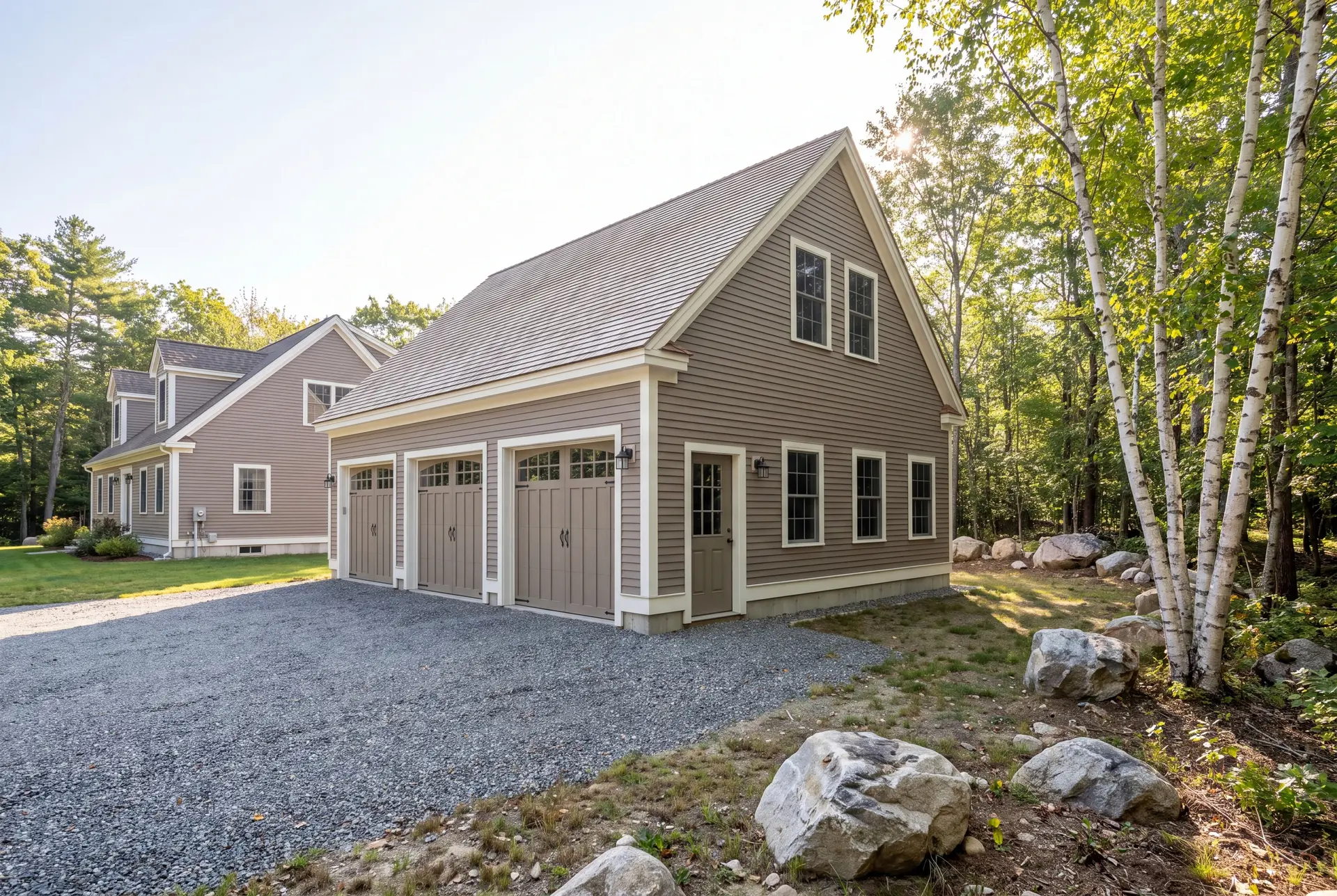 Illustration: A newly built, detached three-bay garage with warm taupe siding and white trim, next to a house, with a gravel driveway and surrounding trees and rocks