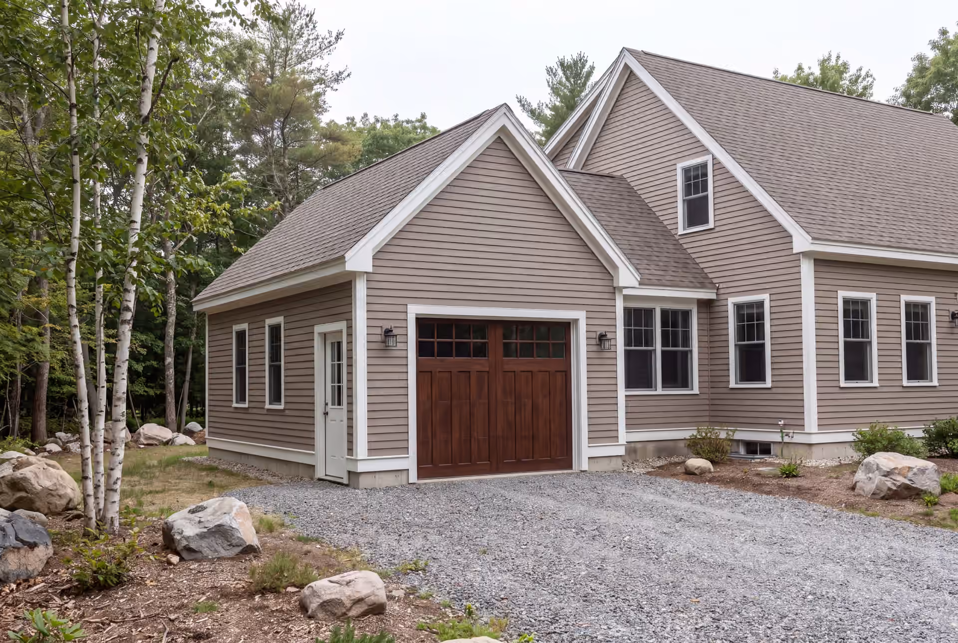 garage builder in Old Orchard Beach, ME: illustrative scene, A newly built, single-bay garage with warm taupe siding and a dark brown carriage-style door, attached to a matching house, with a gravel driveway and trees in the background