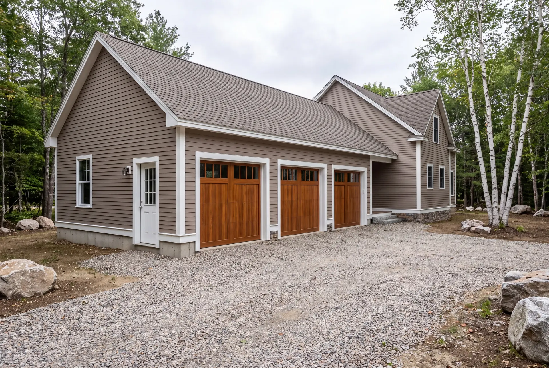 Illustration: A newly built three-bay garage with warm taupe siding and wooden doors, attached to a house, with a gravel driveway and surrounding trees