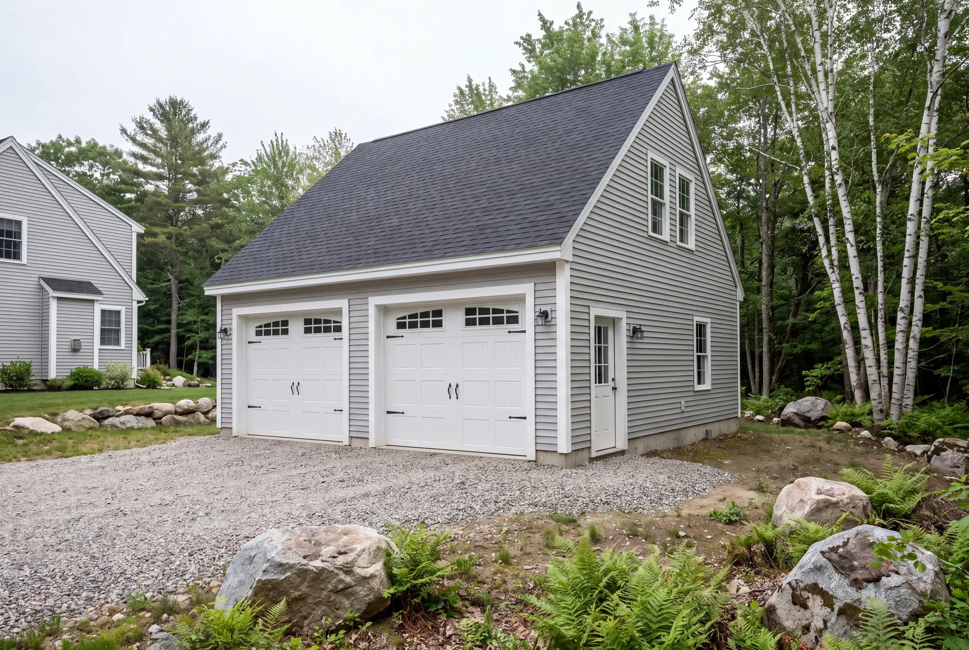 Illustration: A newly built light gray two-car garage with a dark roof and gravel driveway, next to a house and surrounded by trees and rocks