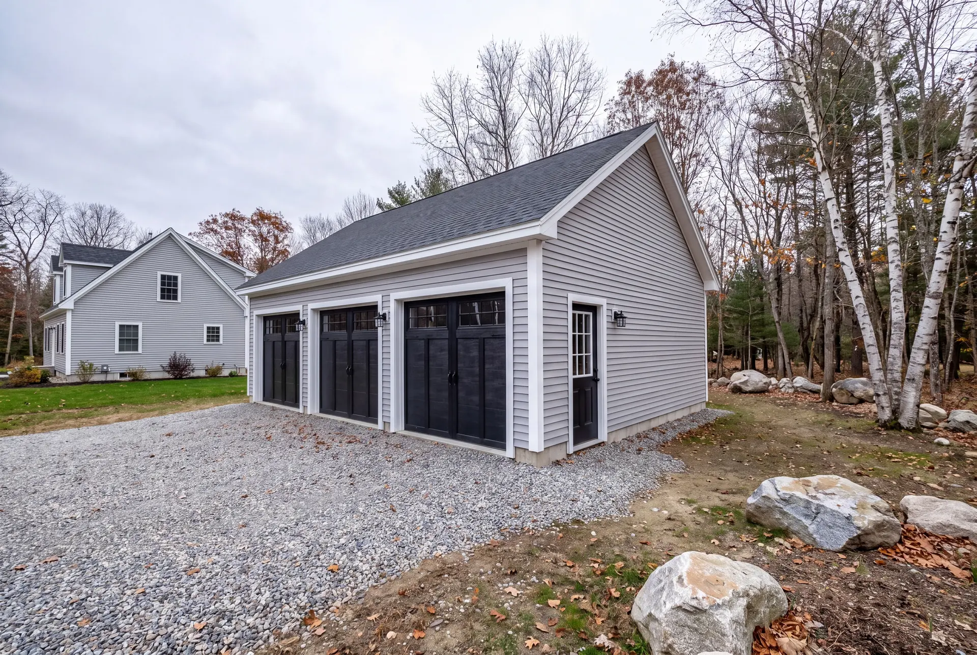 Illustration: A newly built light gray three-bay garage with black doors and a gravel driveway, next to a house and a wooded area with rocks