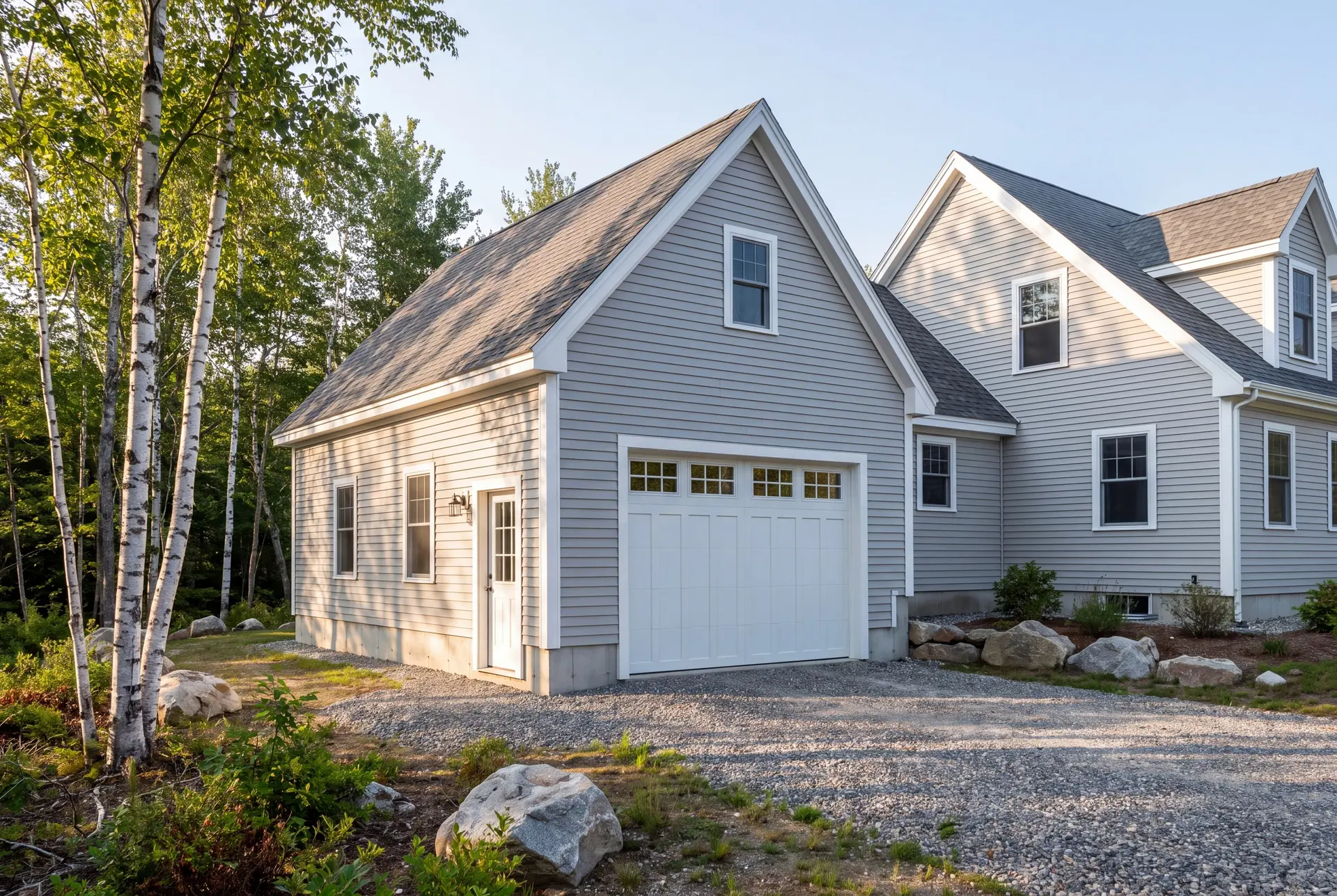 Illustration: A newly built light gray garage with a white carriage door and service door, attached to a matching house, with a gravel driveway and trees