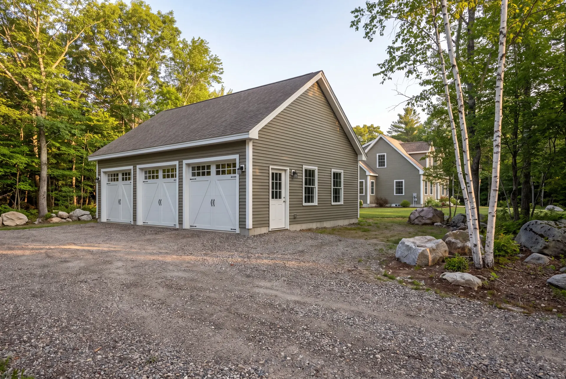Illustration: A newly built three-bay garage with olive siding and white doors, next to a house, surrounded by a gravel driveway, trees, and rocks