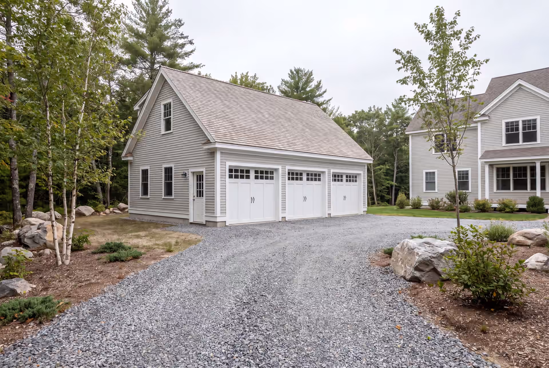 garage builder in Sanford, ME: illustrative scene, A newly built light gray garage with three white doors and a shingle roof, next to a house, with a gravel driveway and trees