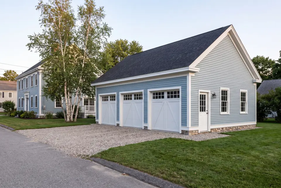 Illustration: A newly built light blue three-car garage with white doors and trim, next to a matching house, with a gravel driveway and green lawn