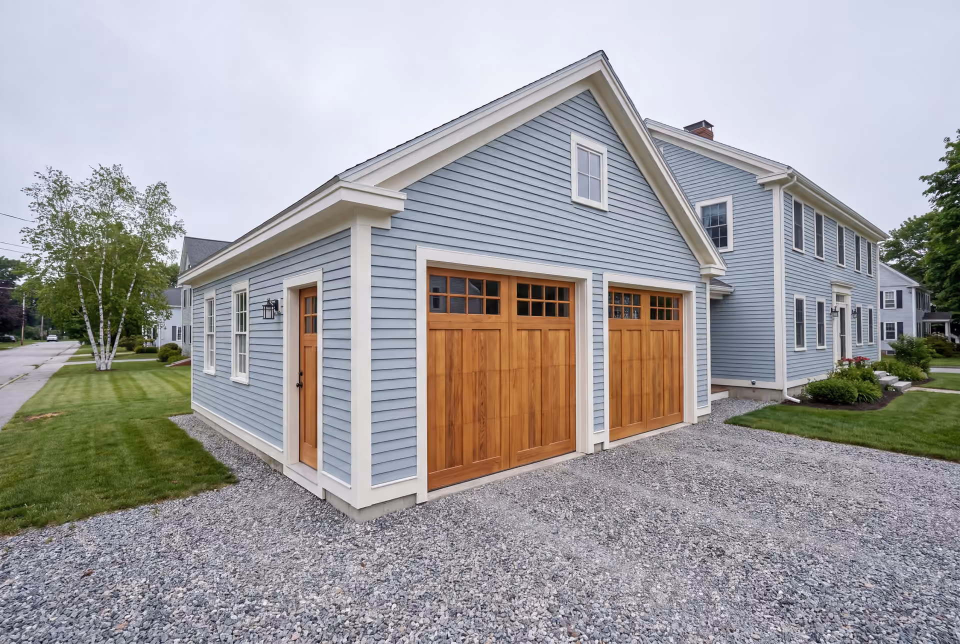 garage builder in South Portland, ME: illustrative scene, A newly built light blue garage with two wooden garage doors and a gravel driveway, next to a matching house