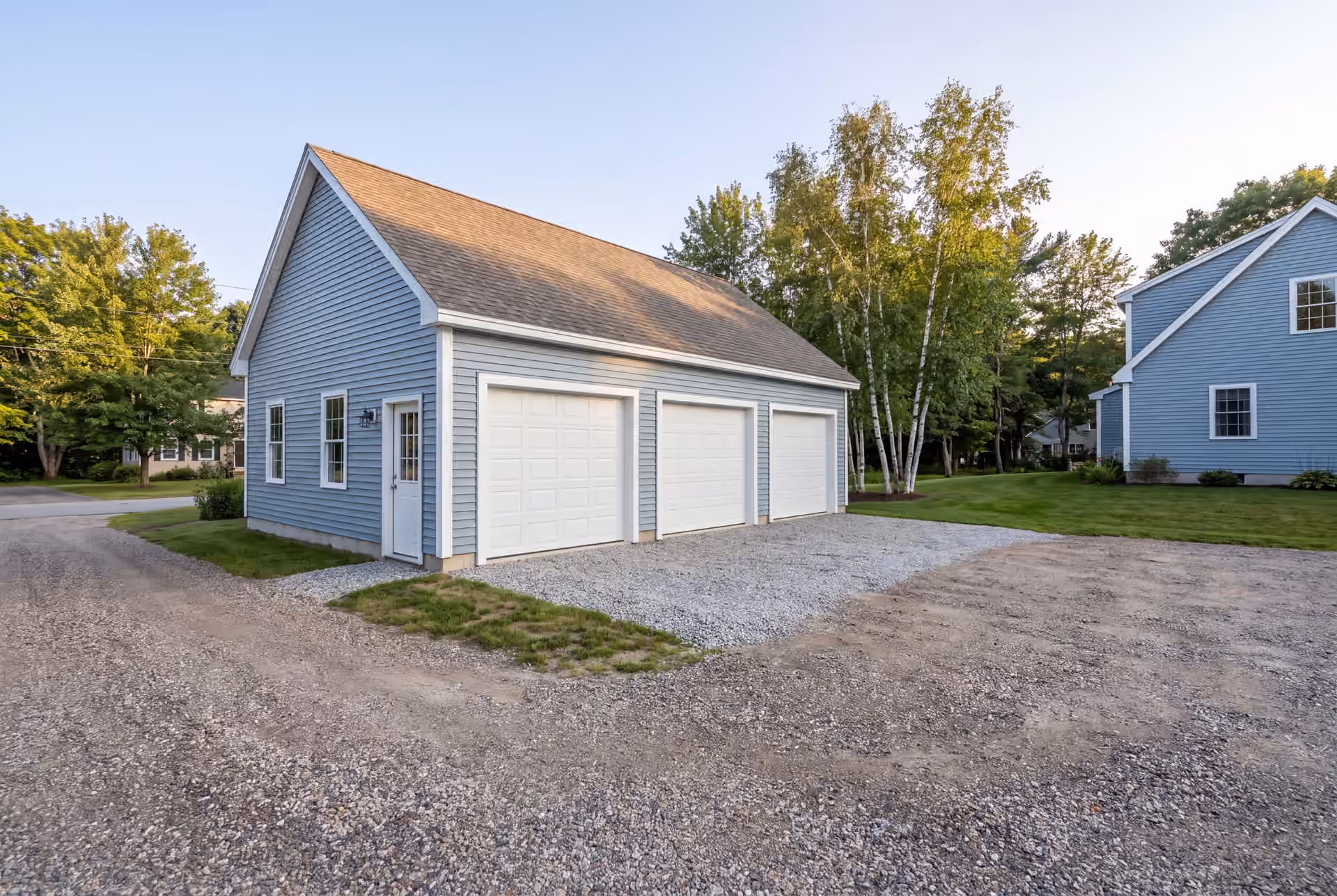 garage builder in Standish, ME: illustrative scene, A newly built light blue garage with three white garage doors and a gravel driveway, next to a matching house